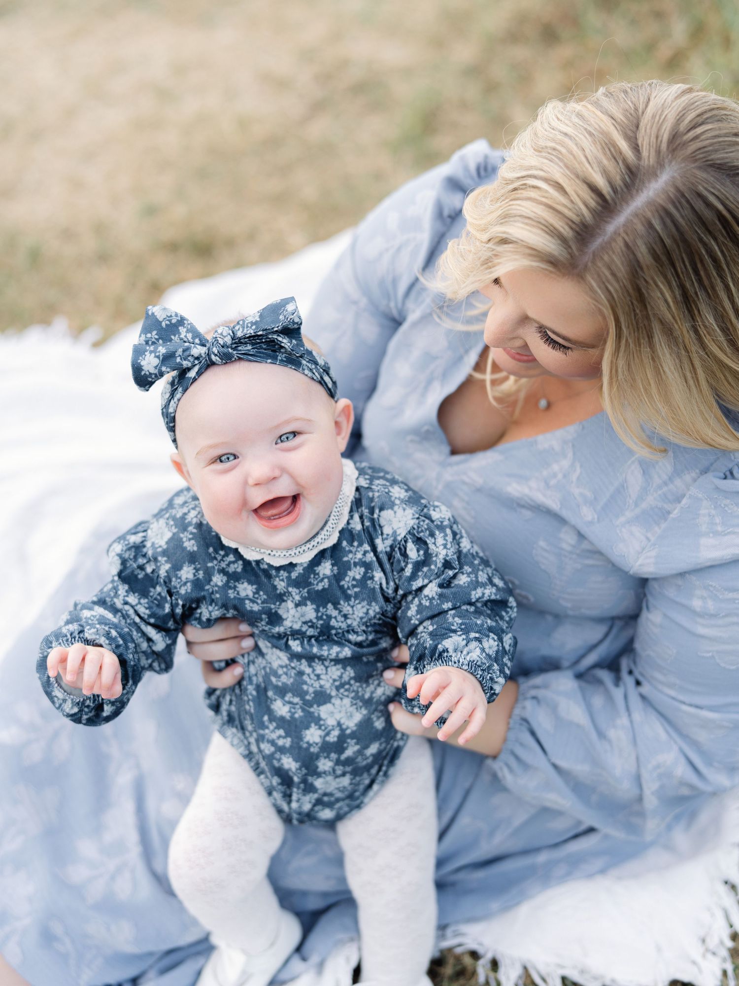 A smiling baby in a floral outfit and bow headband sits on a blanket with a blonde woman in a blue dress after prenatal fitness in Raleigh.