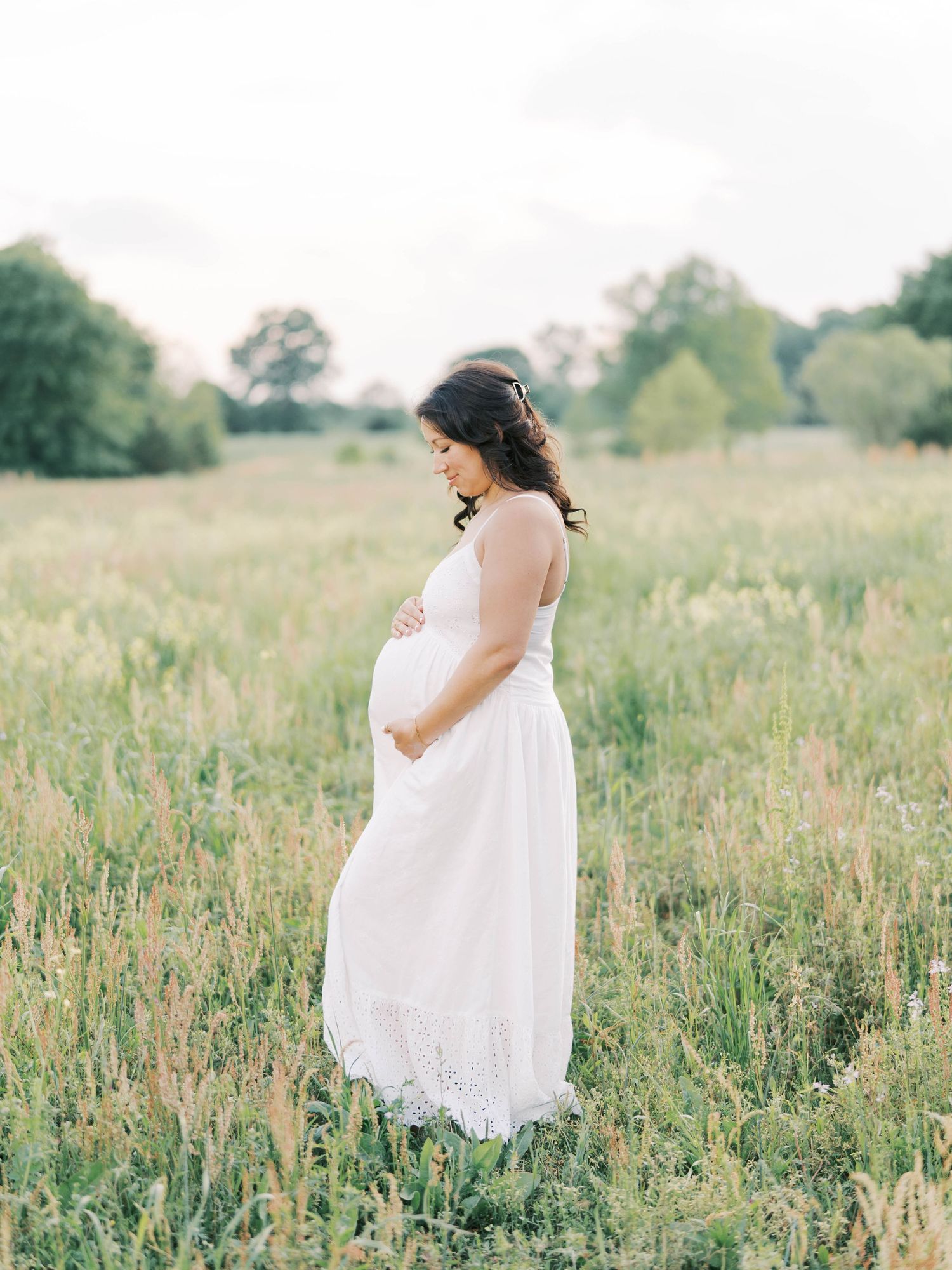 Side profile of expectant mother in white dress standing in wildflower meadow.
