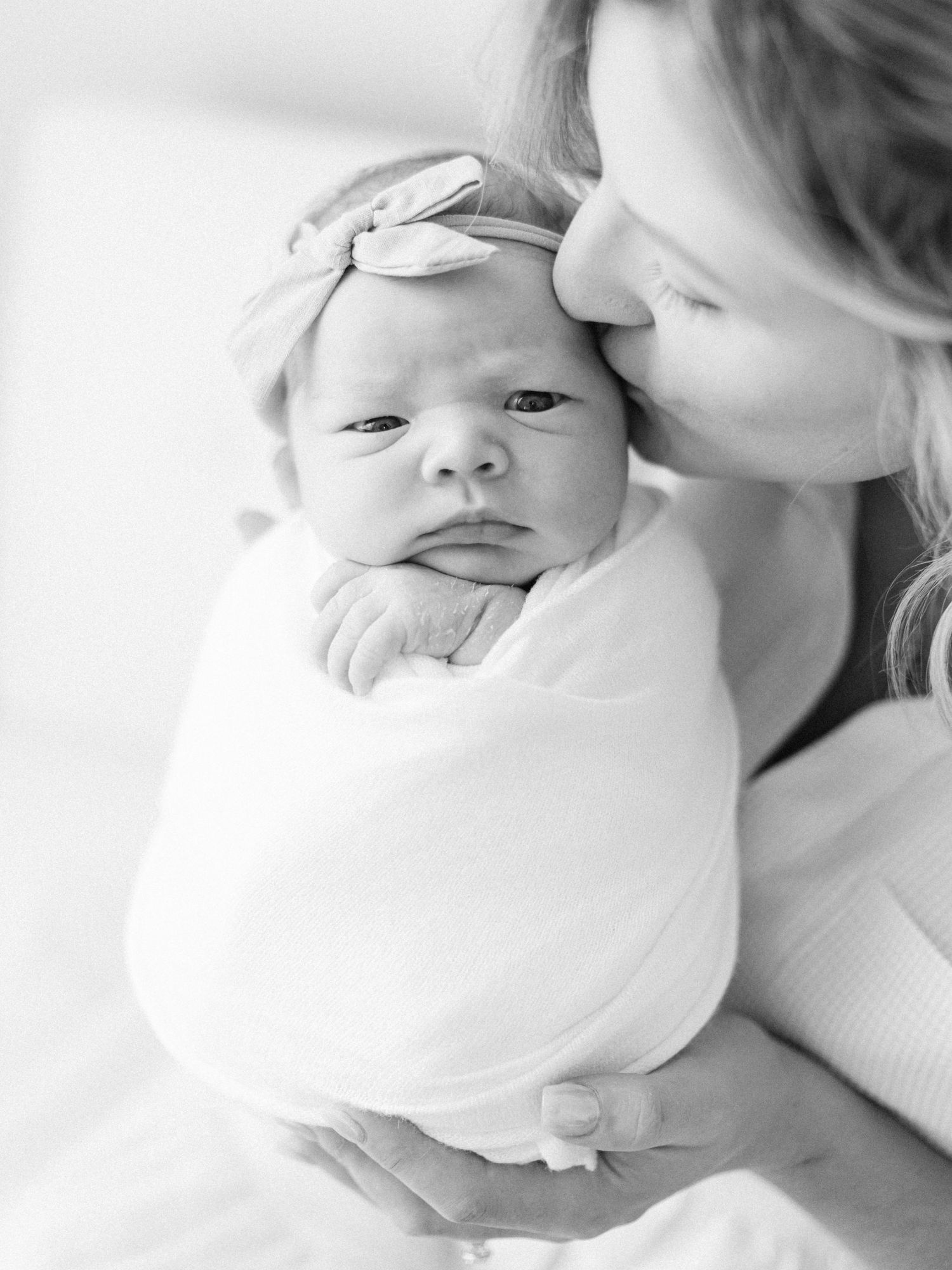 Black and white photo of a mother kissing her swaddled newborn baby wearing a bow headband.