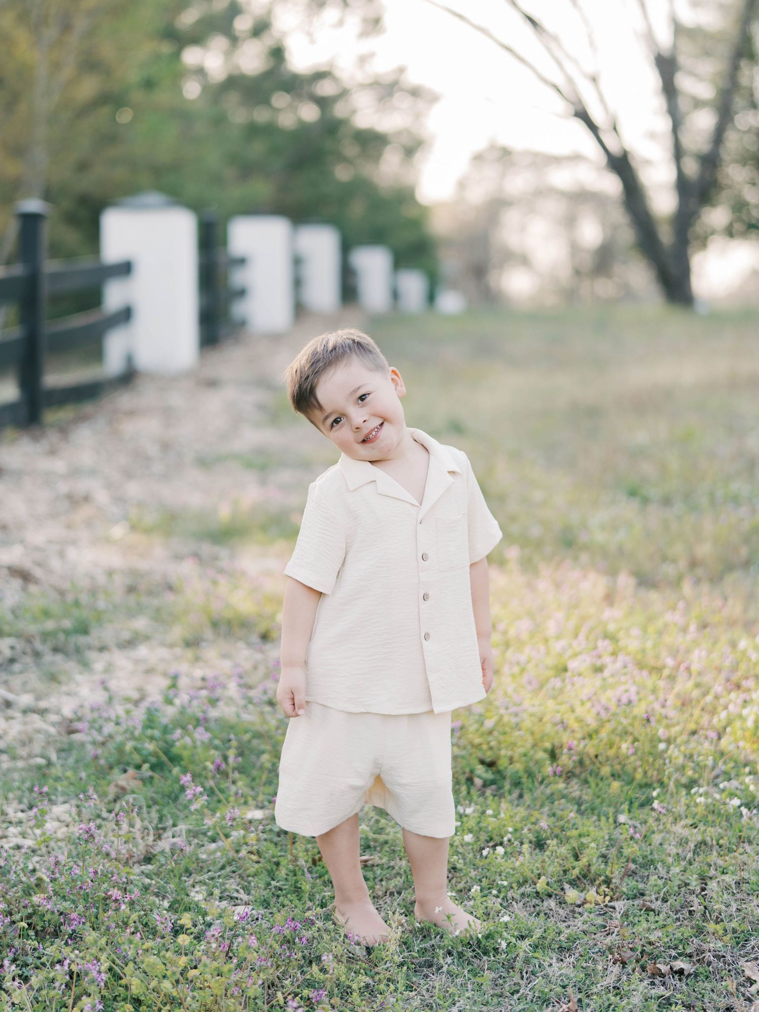 Child in cream-colored outfit playing outdoors near white fence posts at sunset.