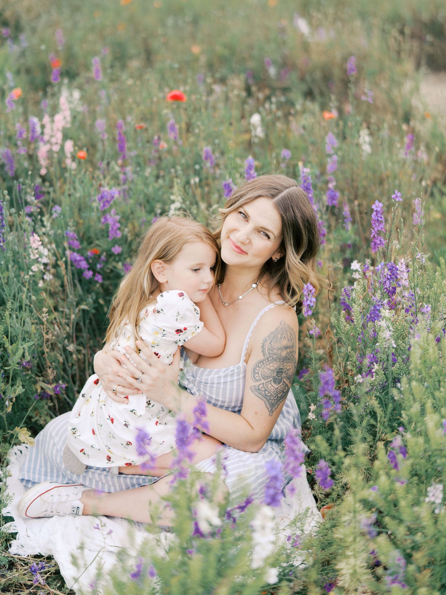 A mother and young daughter embrace in a vibrant wildflower field with purple blooms and poppies.