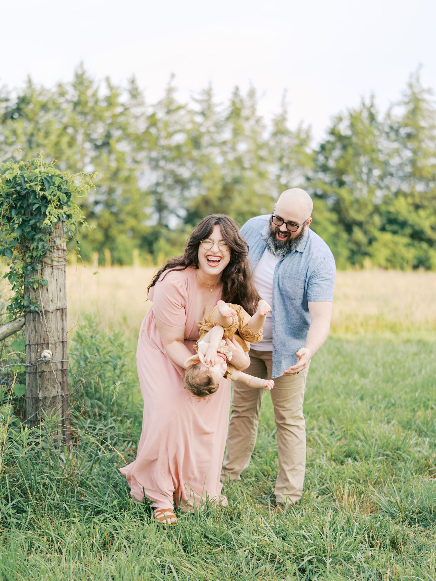 A couple plays with their golden retriever puppy in a sunny meadow surrounded by evergreen trees.