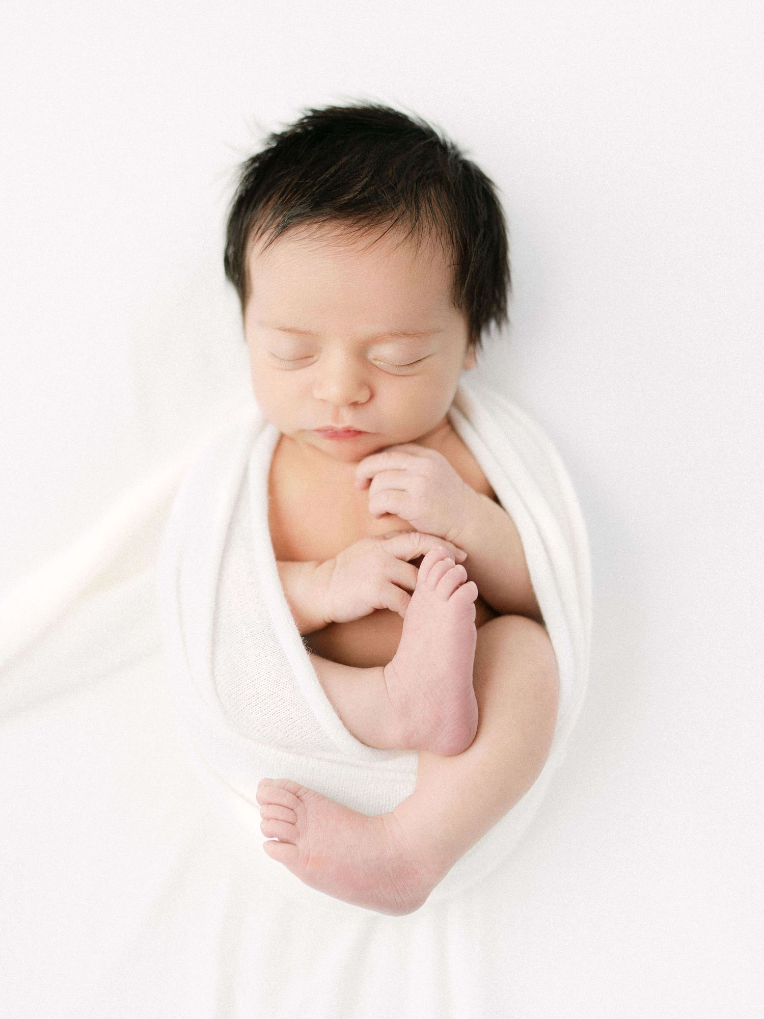 Peaceful newborn baby wrapped in white swaddle blanket, sleeping with tiny feet and hands visible.