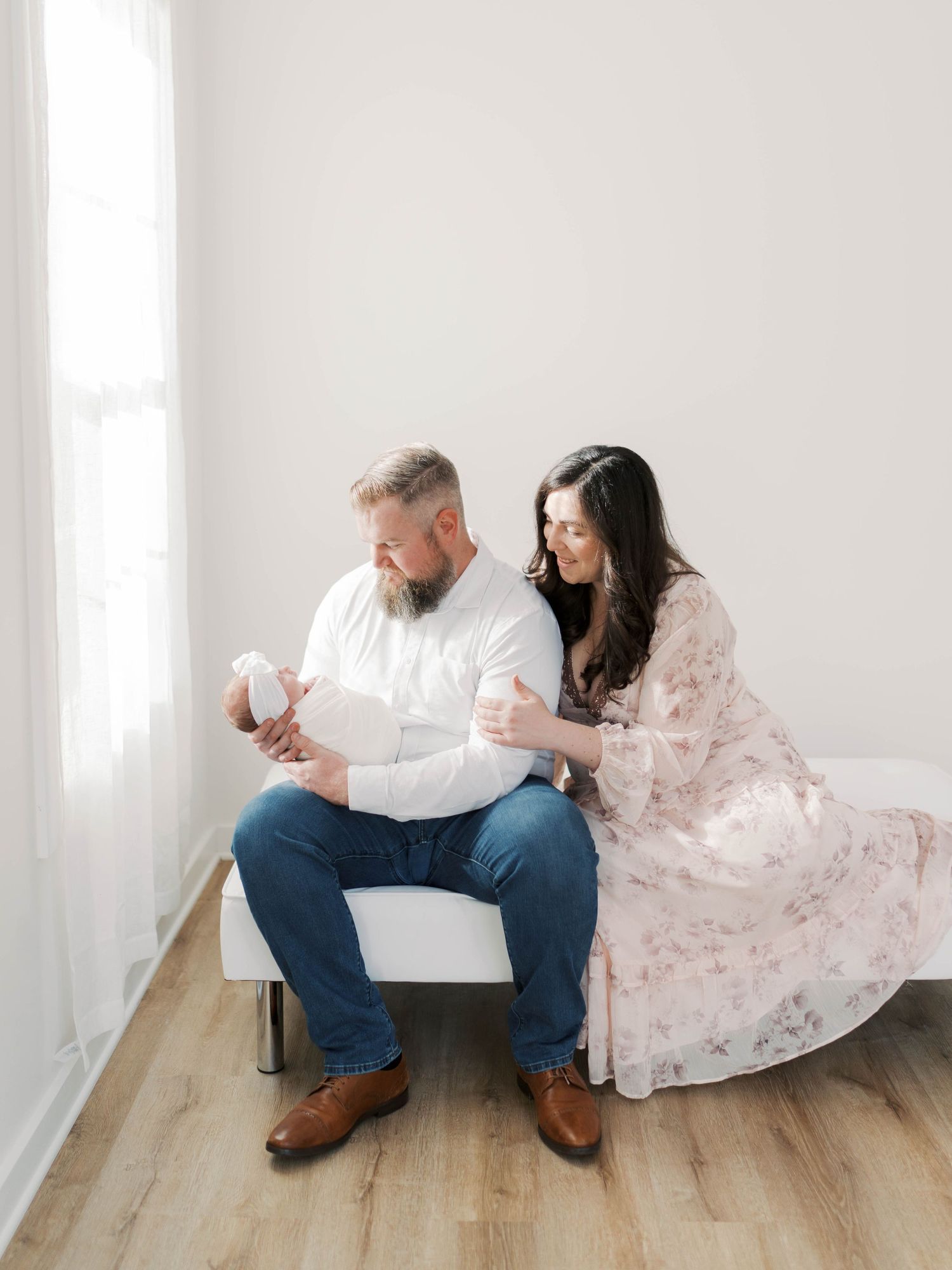 A couple sits on a white bench near a bright window, tenderly gazing at their newborn baby wrapped in white.