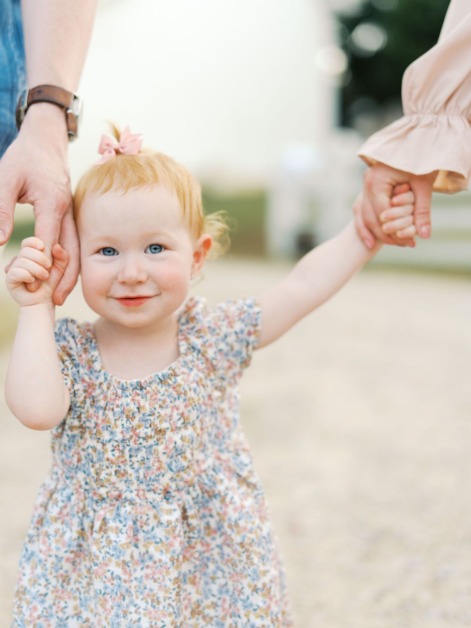 A toddler in a floral dress smiles and holds hands while walking on a sunny day outdoors.