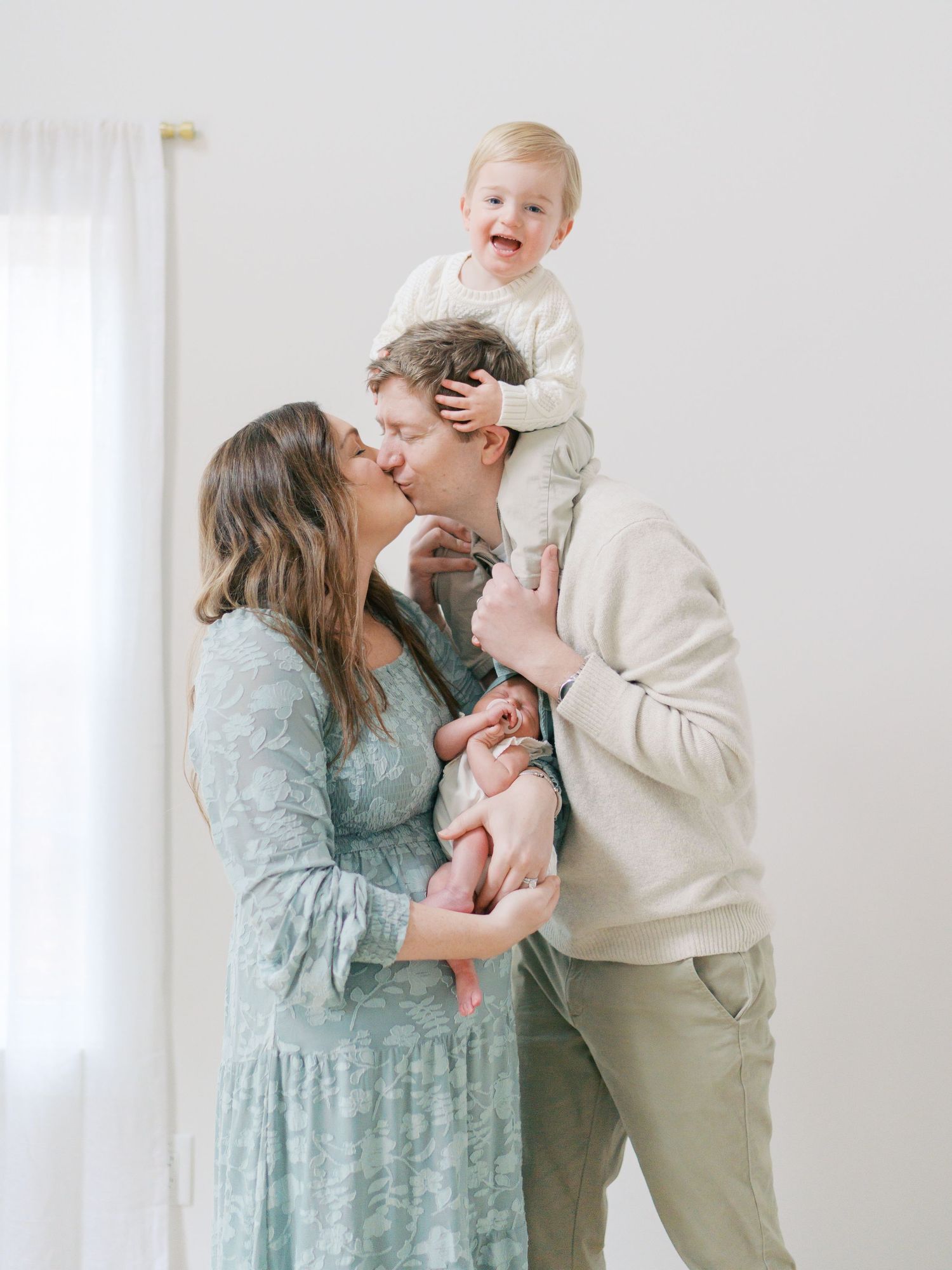 A joyful family of four with parents kissing while dad holds a laughing toddler and mom cradles a newborn baby.
