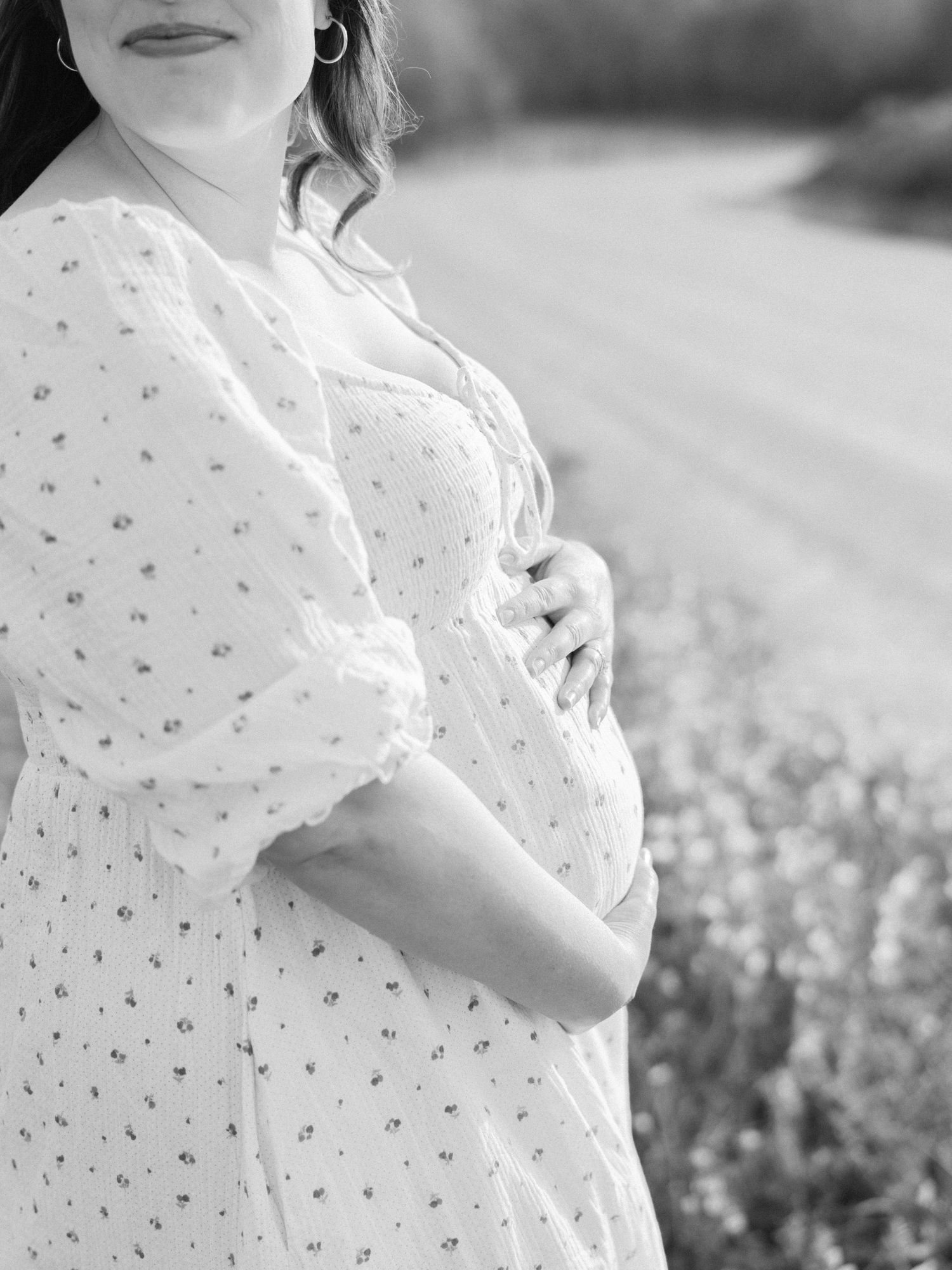 Black and white maternity photo of pregnant woman in floral dress cradling baby bump outdoors.