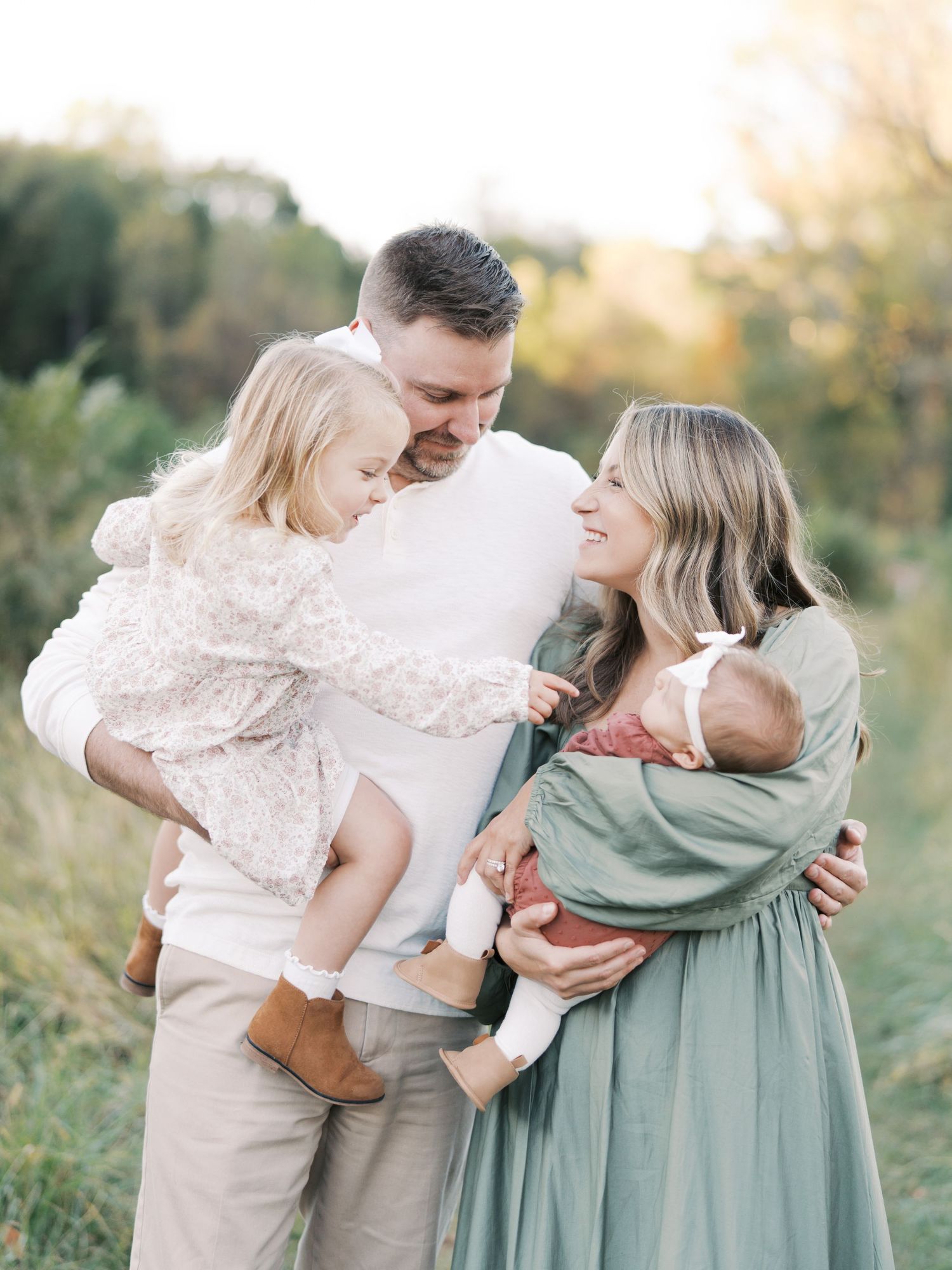 A happy family of four poses outdoors, parents holding a toddler and newborn baby in a sunlit field.