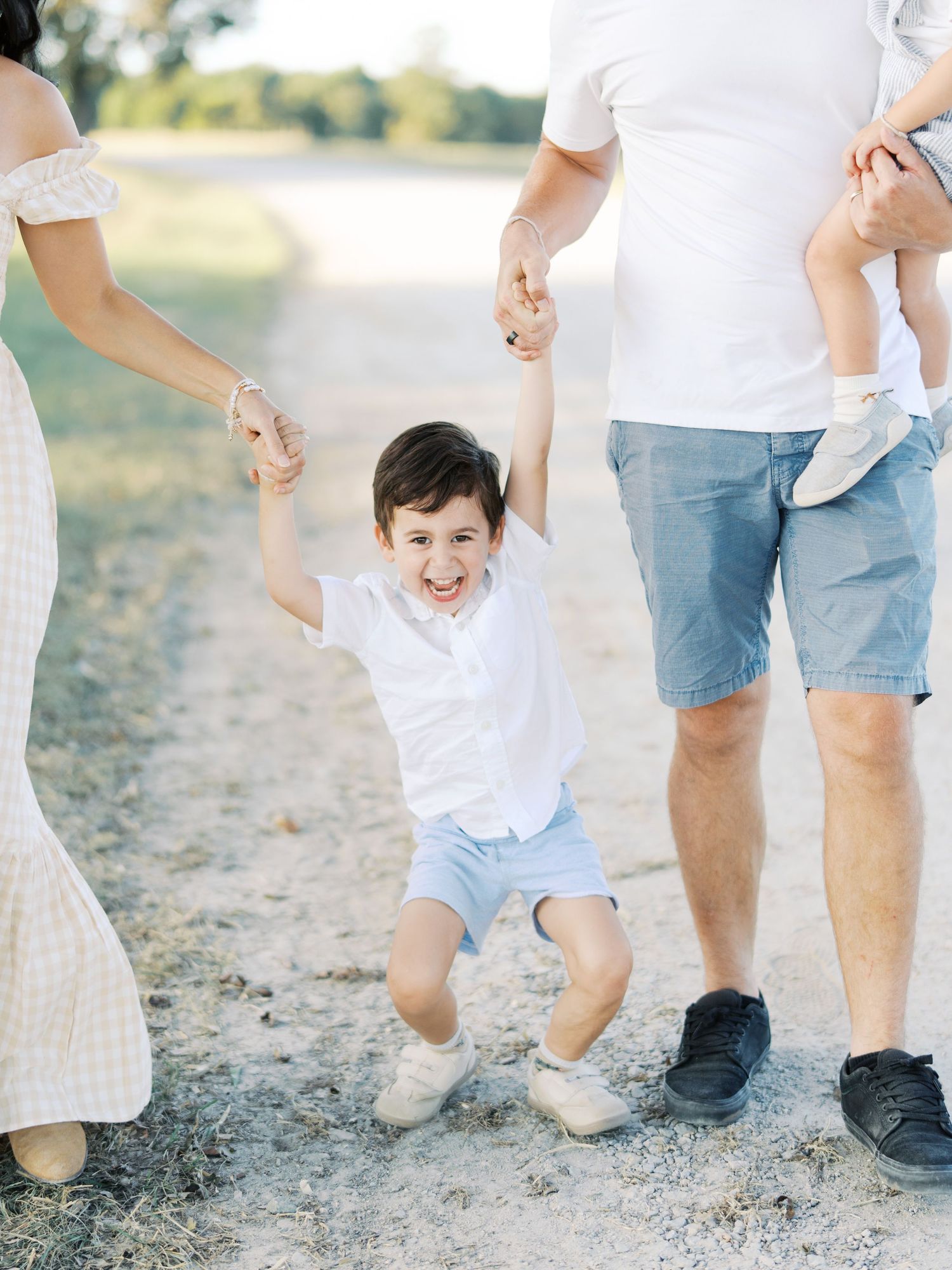 A joyful child in white shirt and blue shorts jumps while holding hands with family members outdoors on a sunny day.
