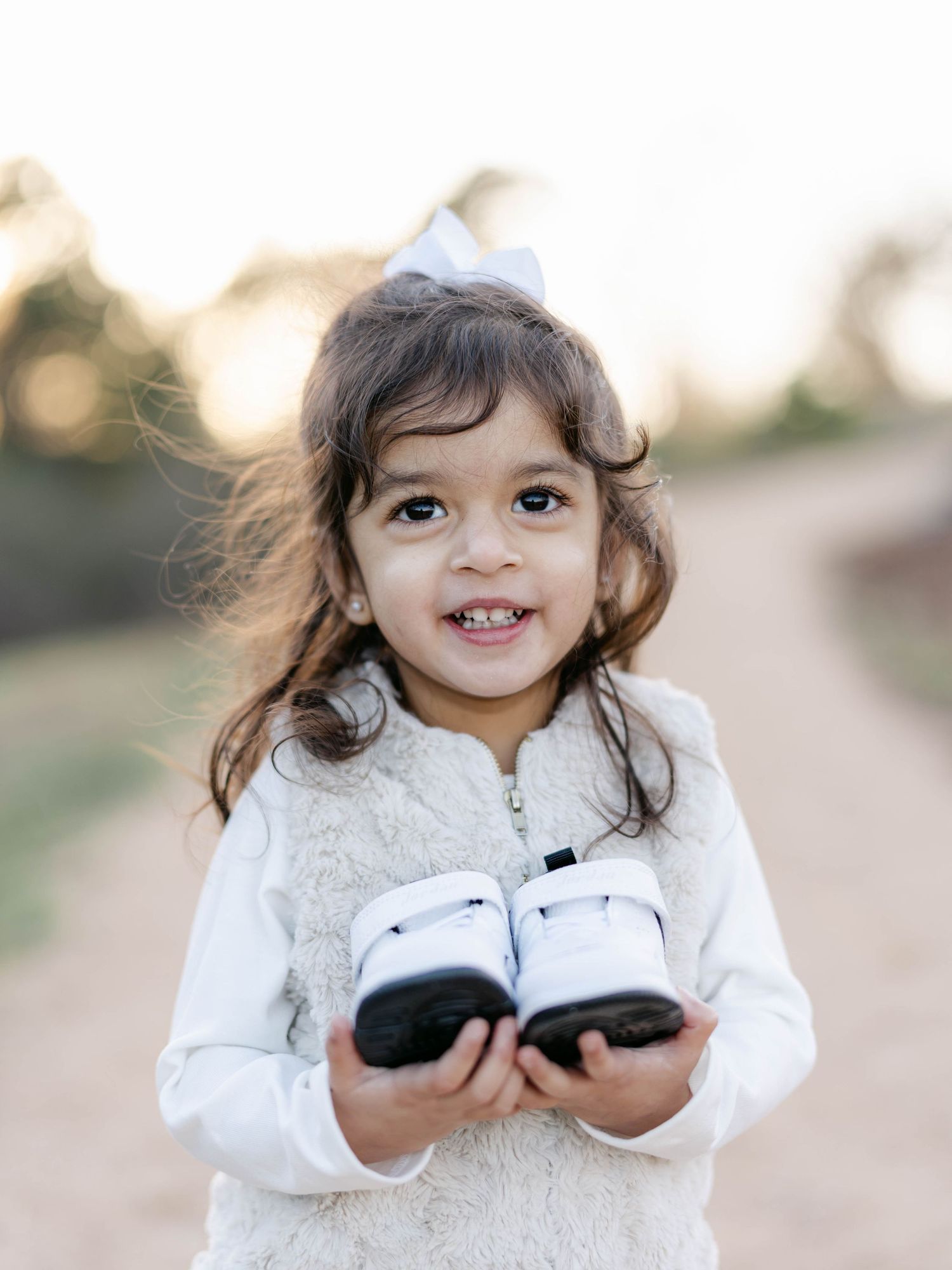 Small child in white sweater smiling while holding pair of baby shoes on outdoor path.