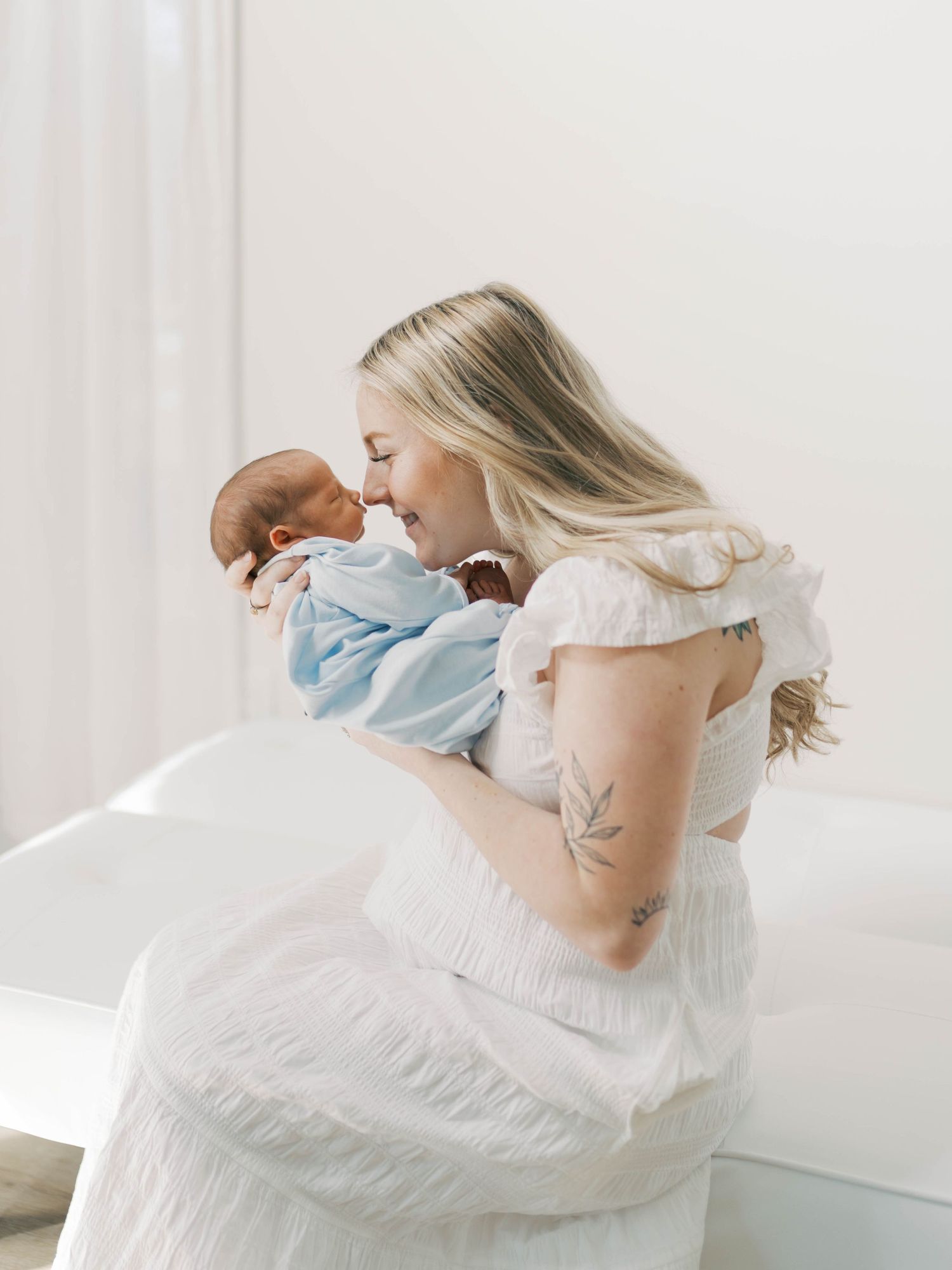 Mother in white dress holding and kissing newborn baby in soft window light.