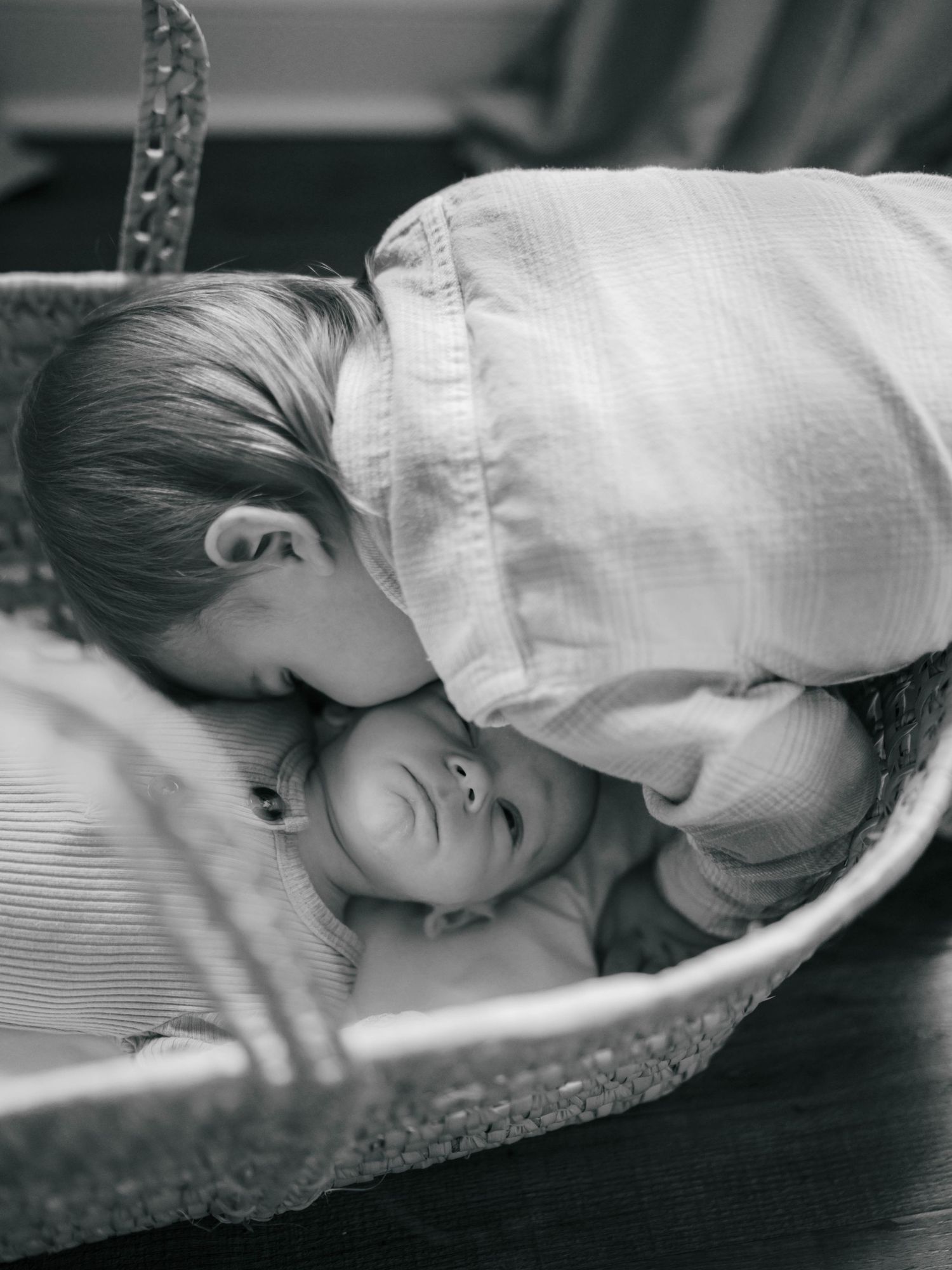 Black and white photo of older child kissing newborn baby lying in a wicker Moses basket.
