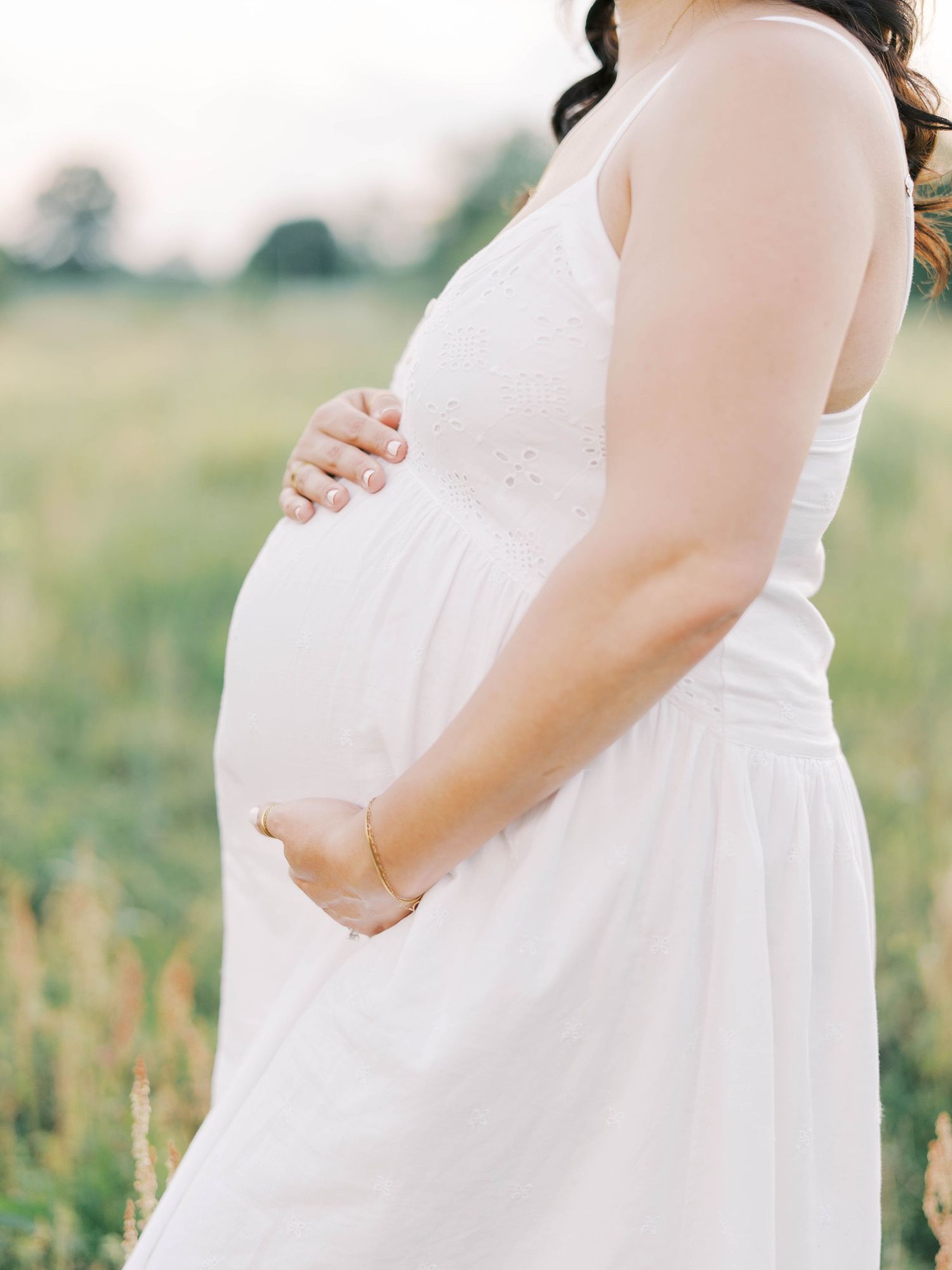 Side view of pregnant woman in white dress tenderly holding her baby bump.