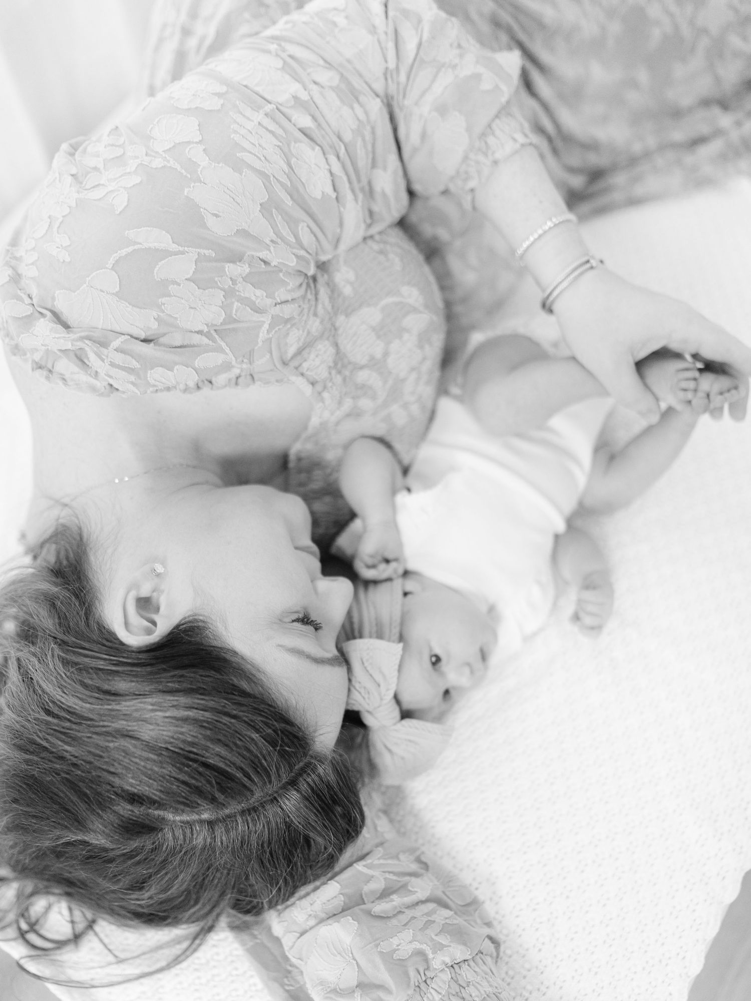 Black and white photo of a mother lying down, tenderly kissing her newborn baby in a soft, intimate moment.
