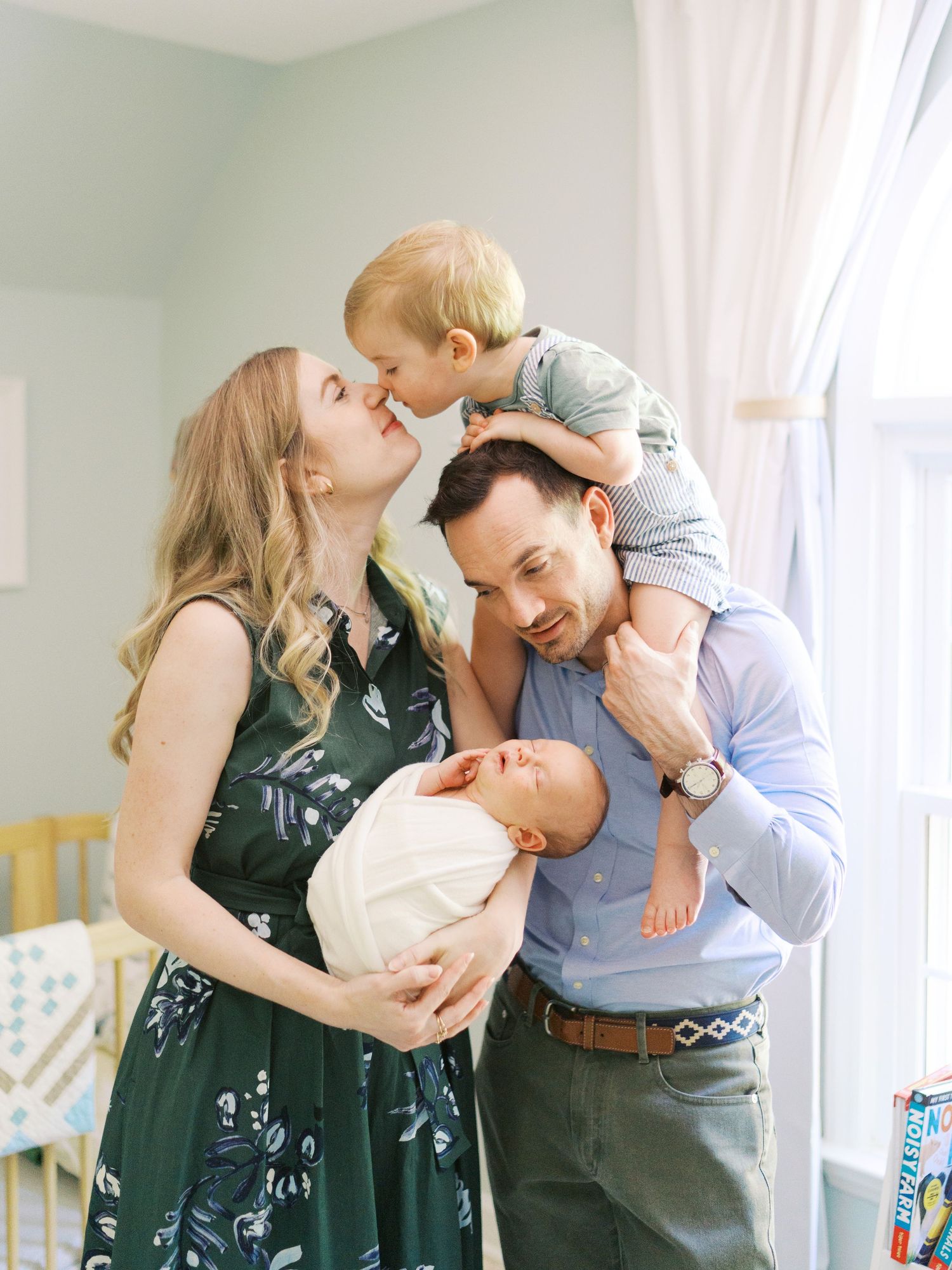 A joyful family portrait with a mother holding a newborn, father with toddler on shoulders, sharing kisses indoors.