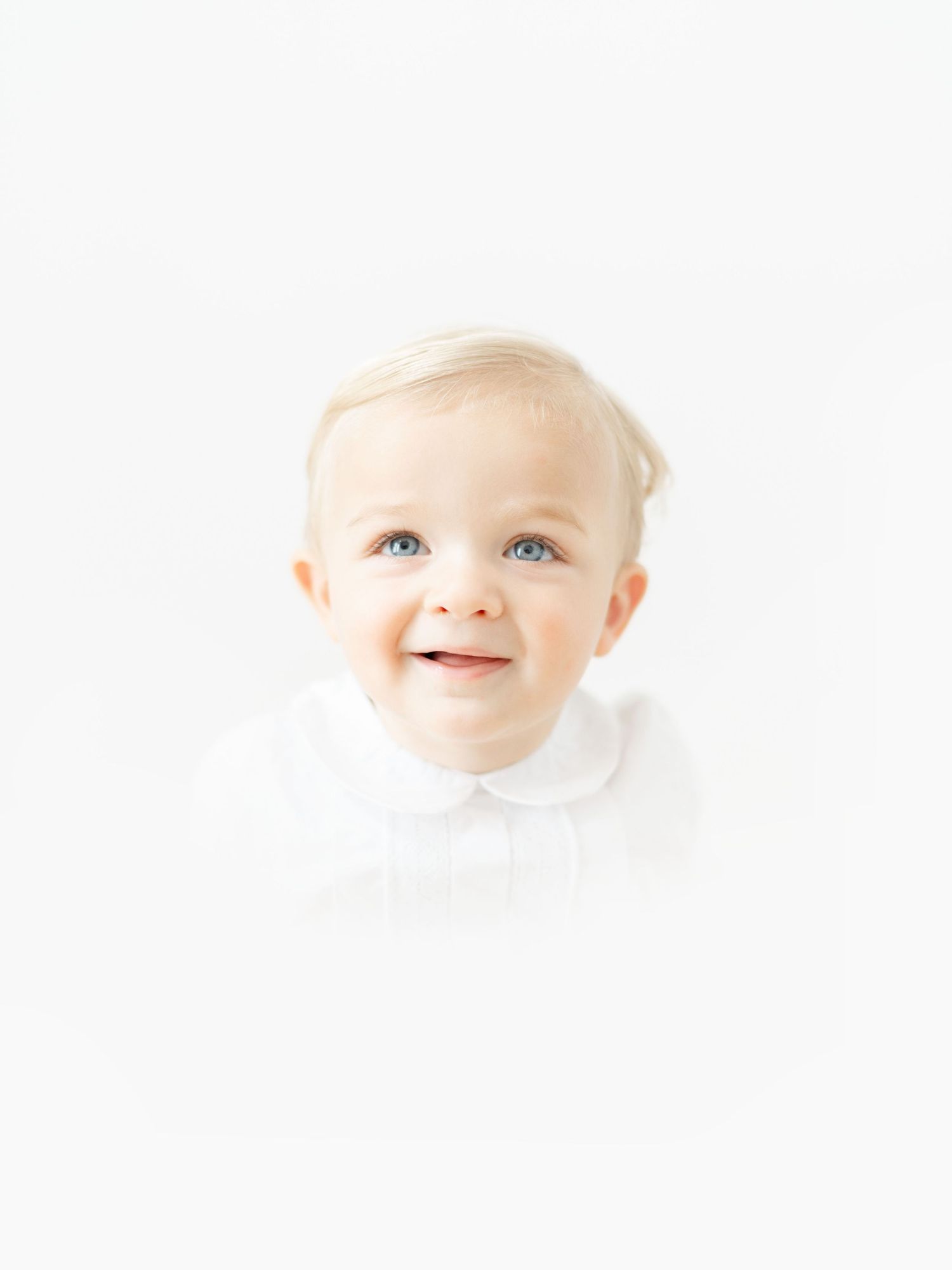 Smiling blonde toddler with blue eyes wearing a white collared shirt against a bright white background.