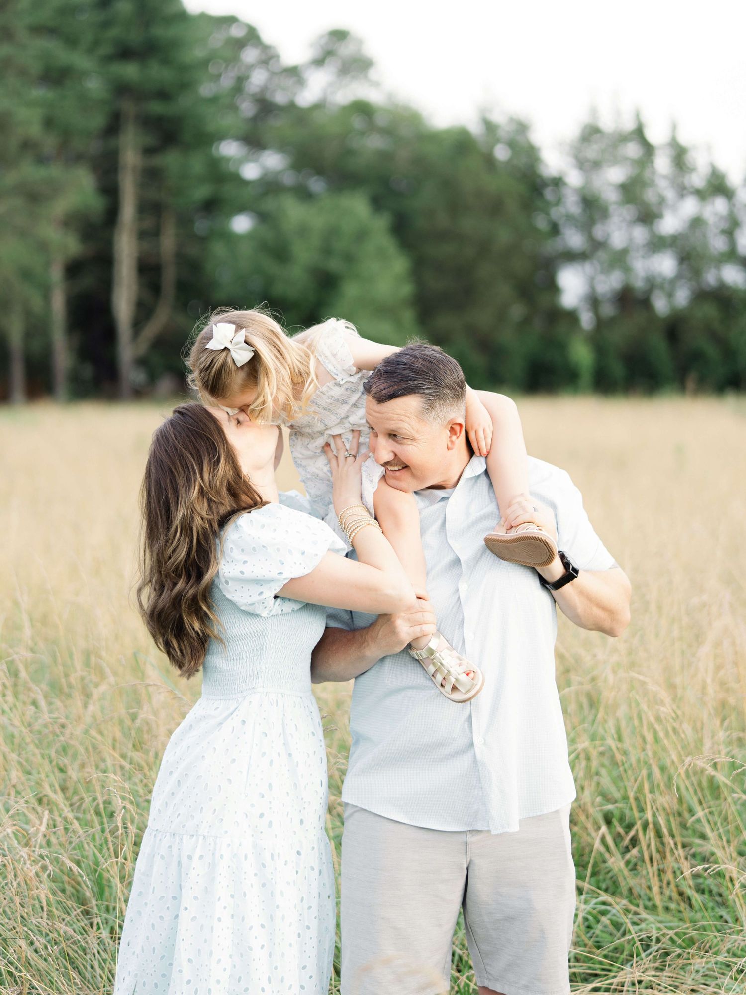 A mom and dad lifting their young daughter in a golden field while they laugh together after visiting pediatric dentists in Raleigh