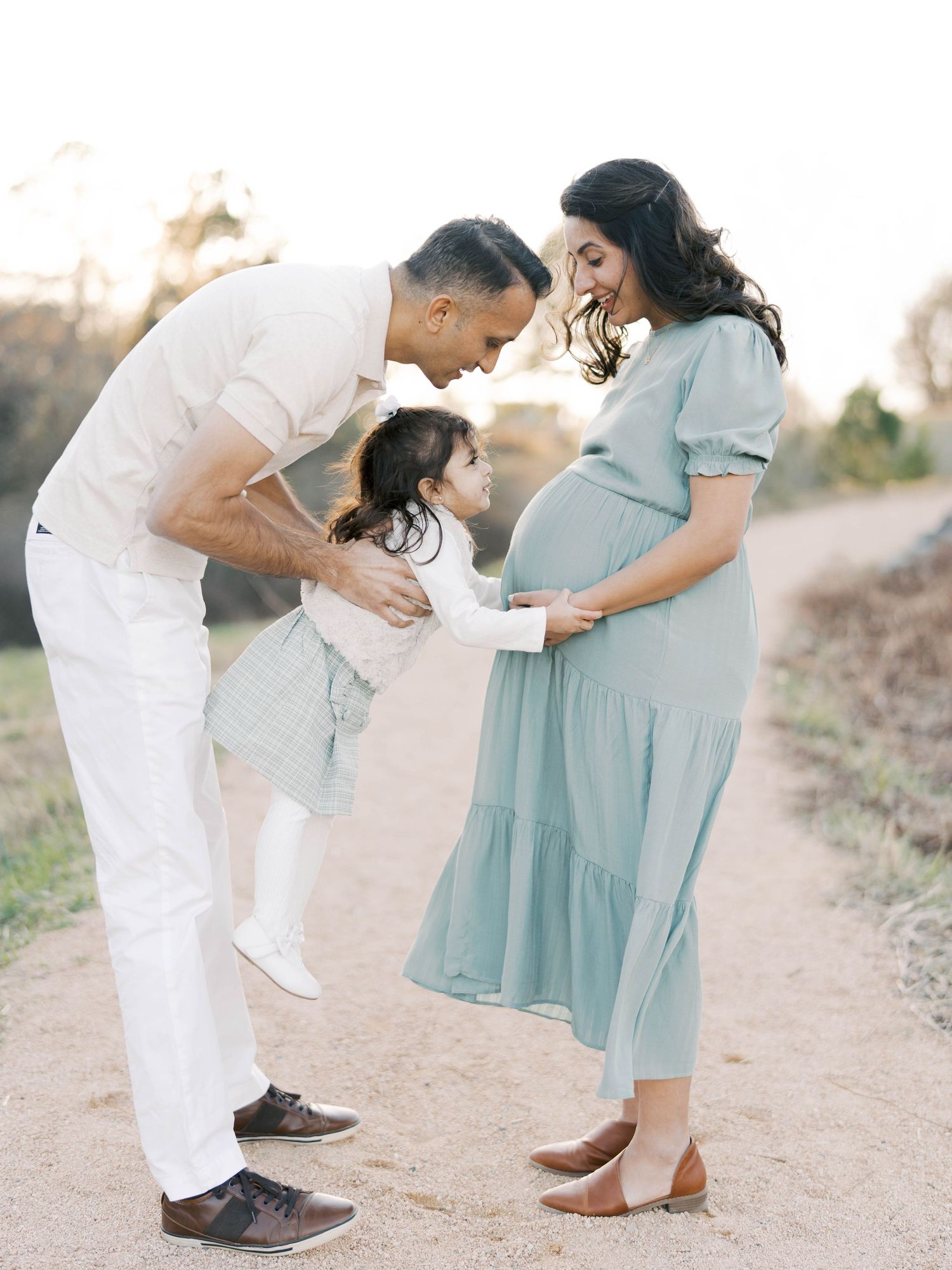 Expecting parents and child on outdoor path looking lovingly at mother's pregnant belly.