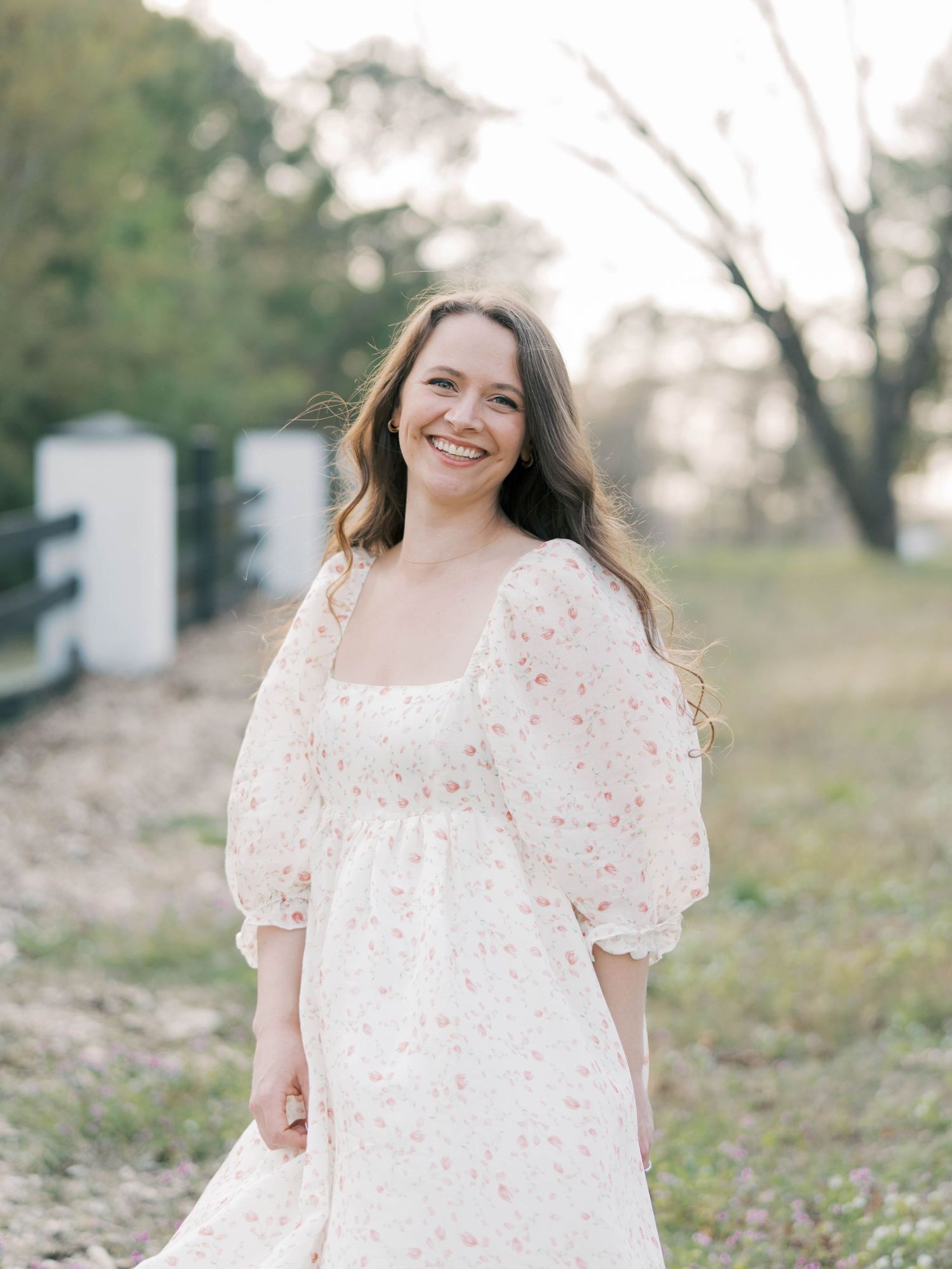 Woman in floral dress smiles while standing near white fence posts at golden hour.