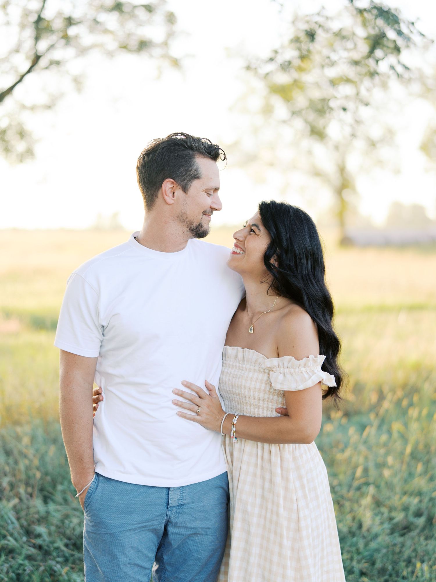 Couple shares intimate moment while laughing together in sunny meadow wearing white and blue casual attire.