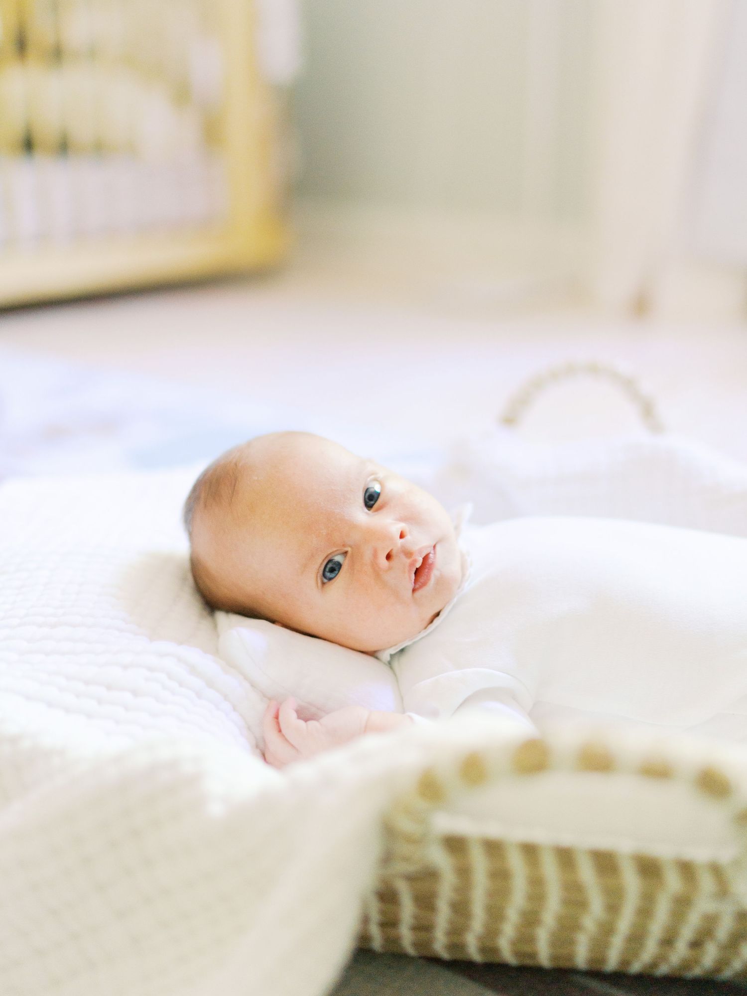 A newborn baby with blue eyes lying in a woven Moses basket on a white knit blanket in a nursery.