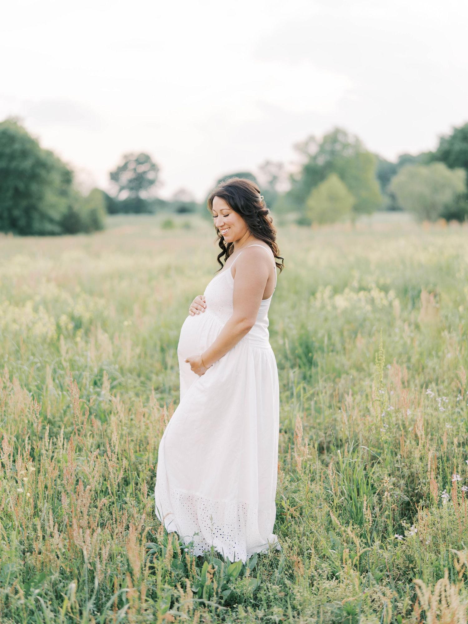 Pregnant woman in a white dress standing in a field, cradling her baby bump during a maternity session after a prenatal massage in Raleigh