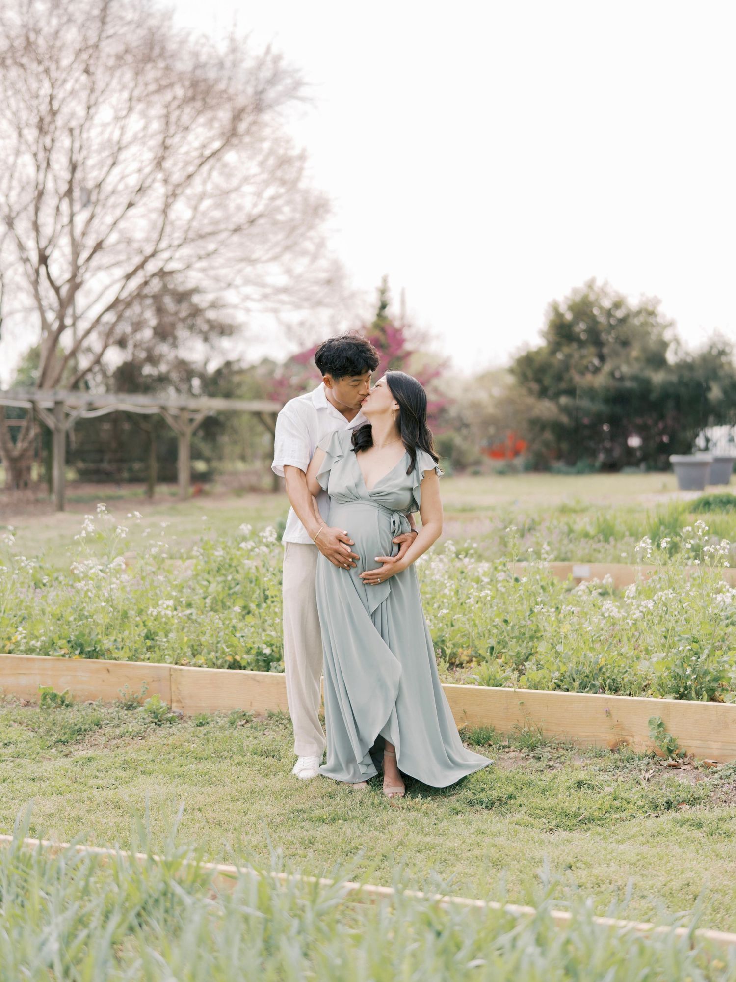 Romantic maternity photo of couple in light-colored outfits standing in garden setting.