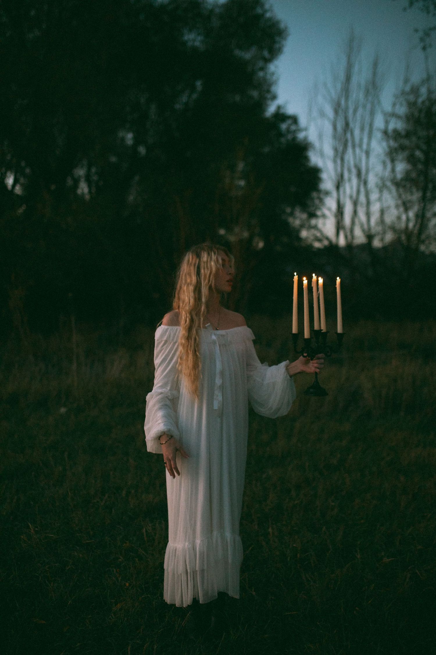 Woman in a flowing white nightgown holds a lit candelabra in a dim grassy field at dusk, with dark trees behind her.