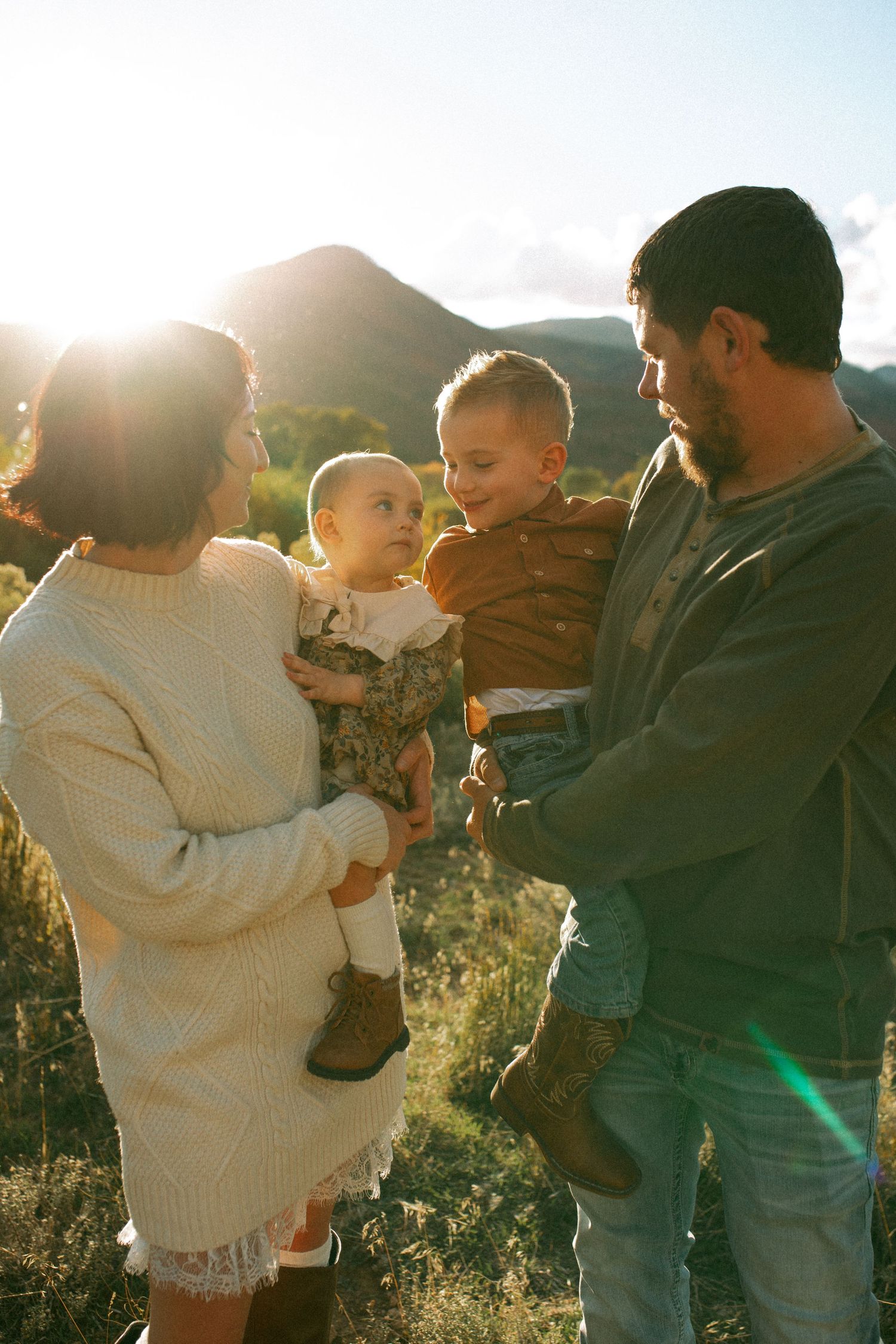 Family of four in golden sunset light, parents holding two young children in front of mountains and grassy hills.
