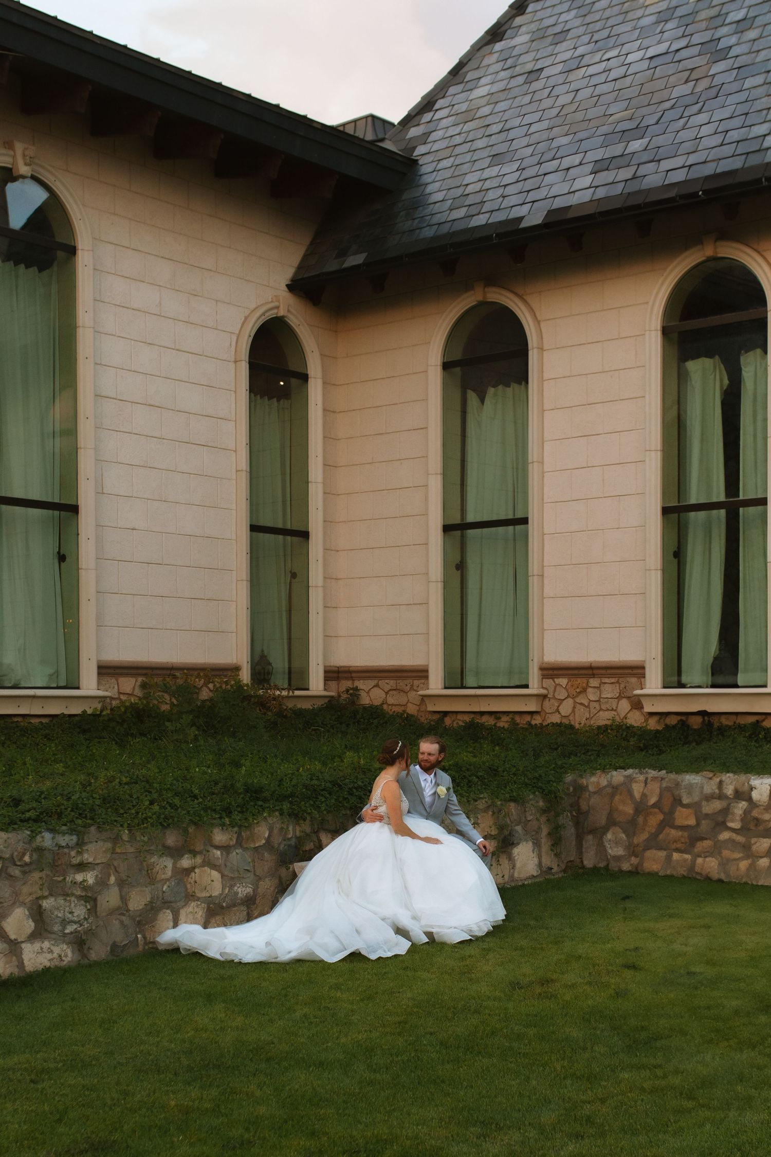 Bride in a flowing white gown and groom in a gray suit sit together on a stone wall in front of an elegant building with arched windows.