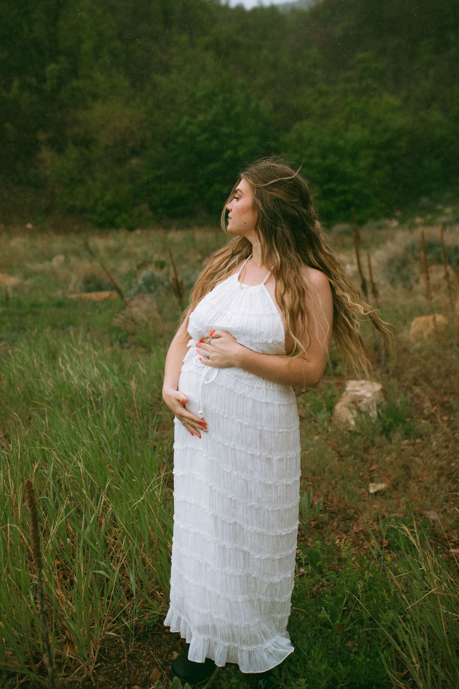 Pregnant woman in a white ruffled dress cradles her baby bump while standing in a grassy field with green hills behind her.