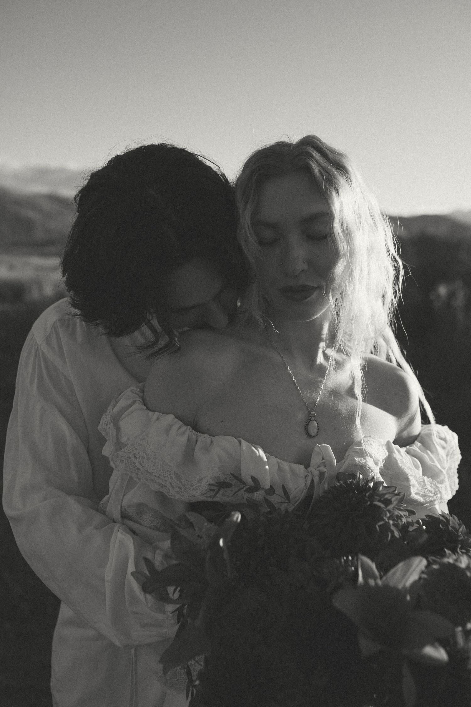 Black and white portrait of a dark-haired man embracing a blonde woman in an off-shoulder lace dress holding a bouquet outdoors.