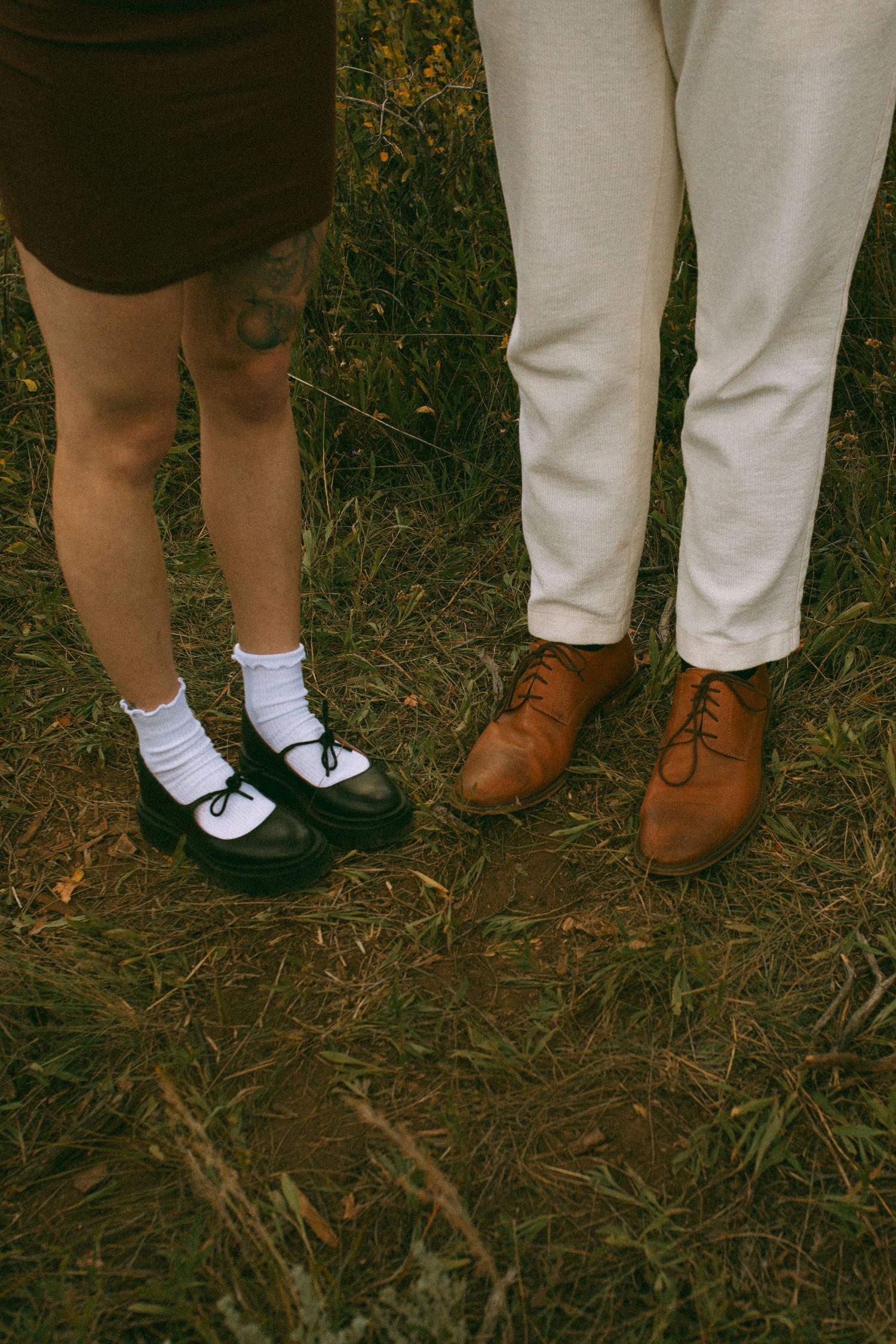 Couple standing close on a grassy field, one in white socks and black Mary Jane shoes, the other in cream trousers and brown leather shoes.