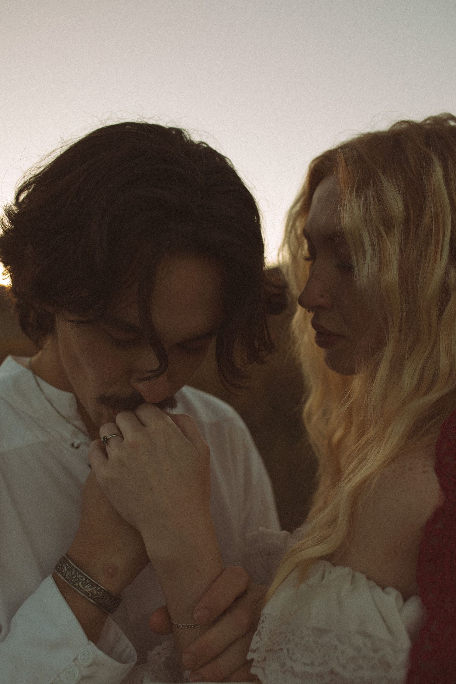A dark-haired man in a white shirt kisses the hand of a blonde woman in a lace-trimmed top during a moody, golden-hour couple portrait.