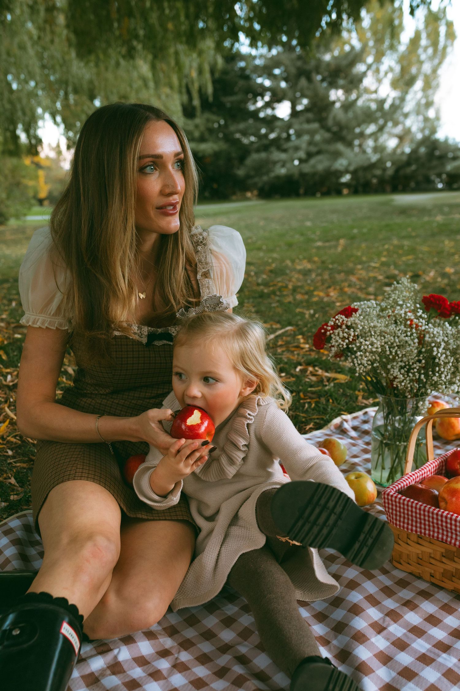 Woman in a puffed-sleeve top and toddler in a beige dress eating a red apple on a checkered picnic blanket in a sunny autumn park.