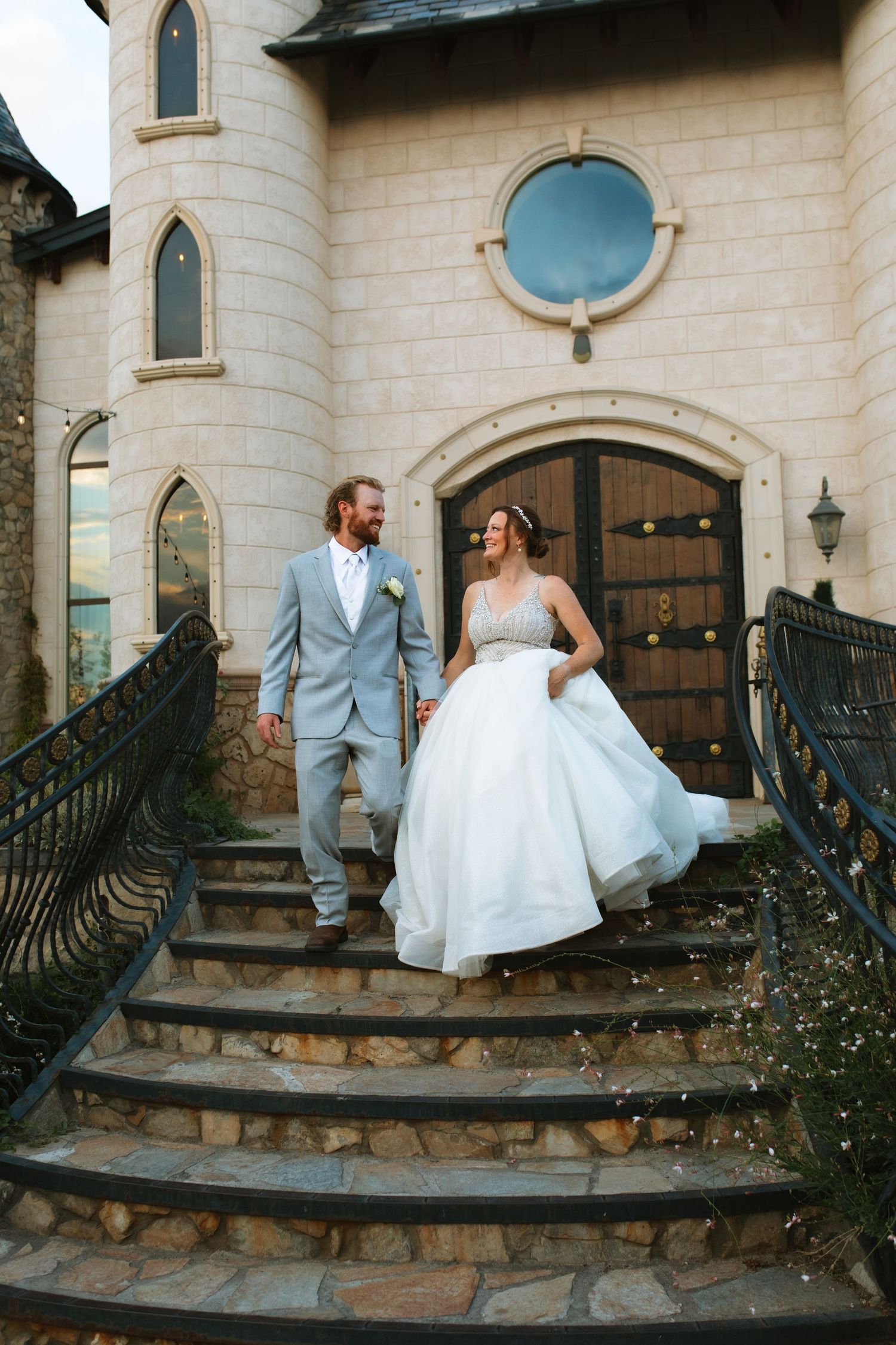 Bride in a white ball gown and groom in a light gray suit walk hand in hand down stone steps outside a castle-style wedding venue.