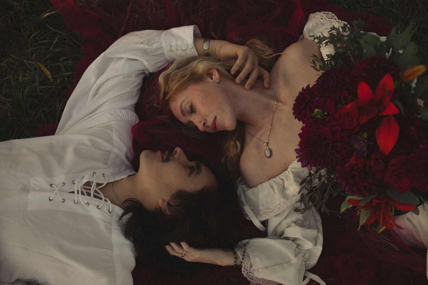 Two people in white romantic blouses lie side by side on dark red flower petals, with a bouquet of deep red lilies and roses beside them.