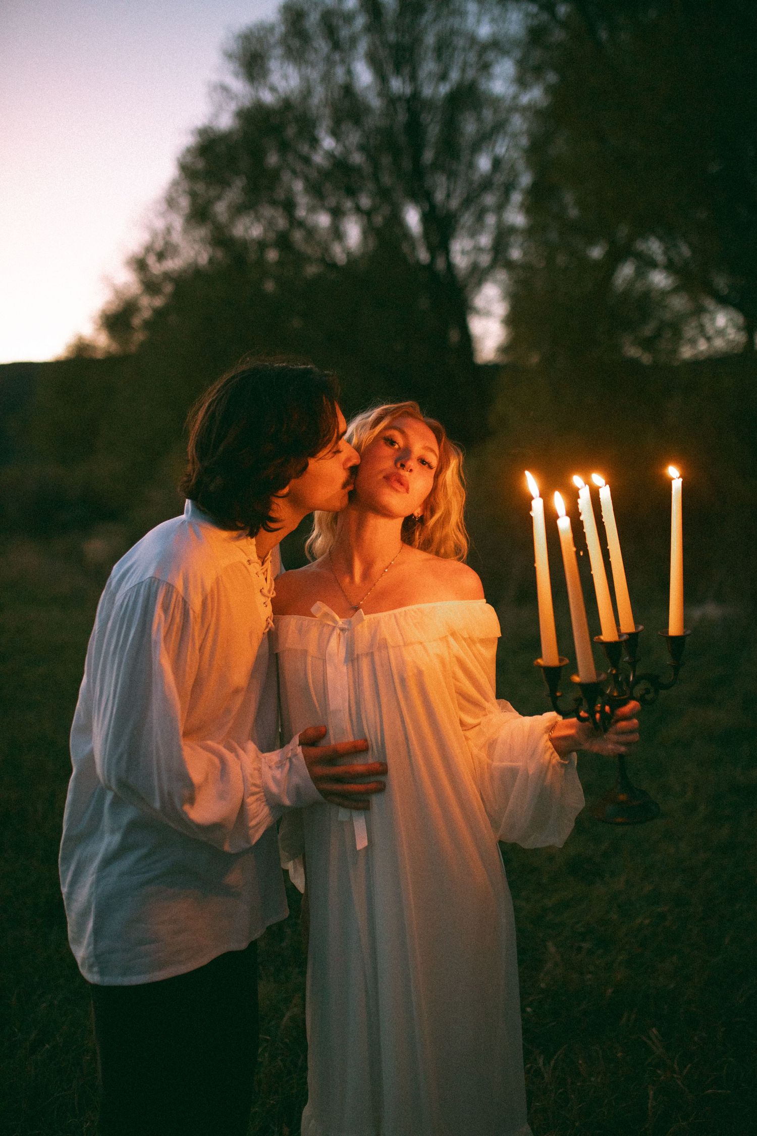 Romantic couple in white vintage clothing share a kiss while holding a lit candelabra outdoors in a grassy field at dusk.