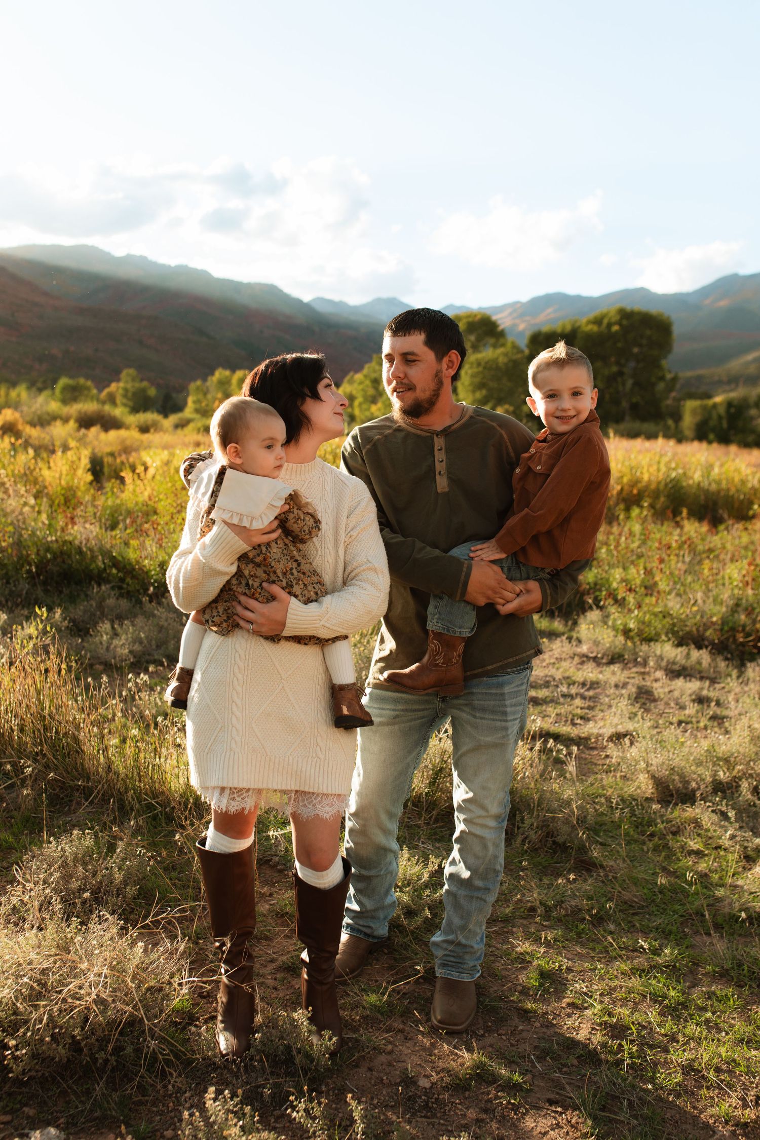 Family of four in warm autumn outfits standing together in a golden grassy field with mountains and soft sunset light behind them.