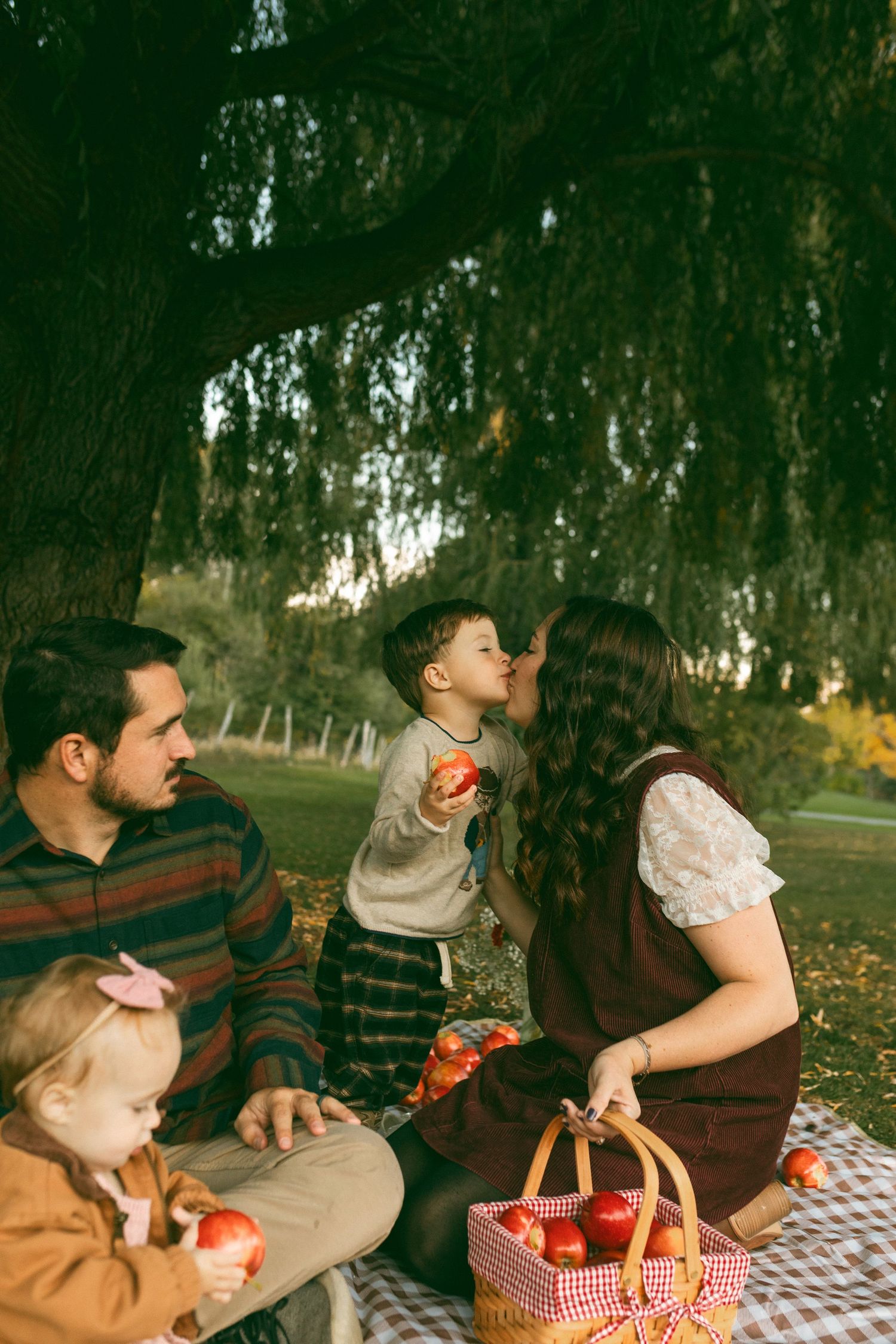 Family enjoys an autumn apple picnic under a willow tree as a mother kisses her toddler son holding a red apple beside a basket.