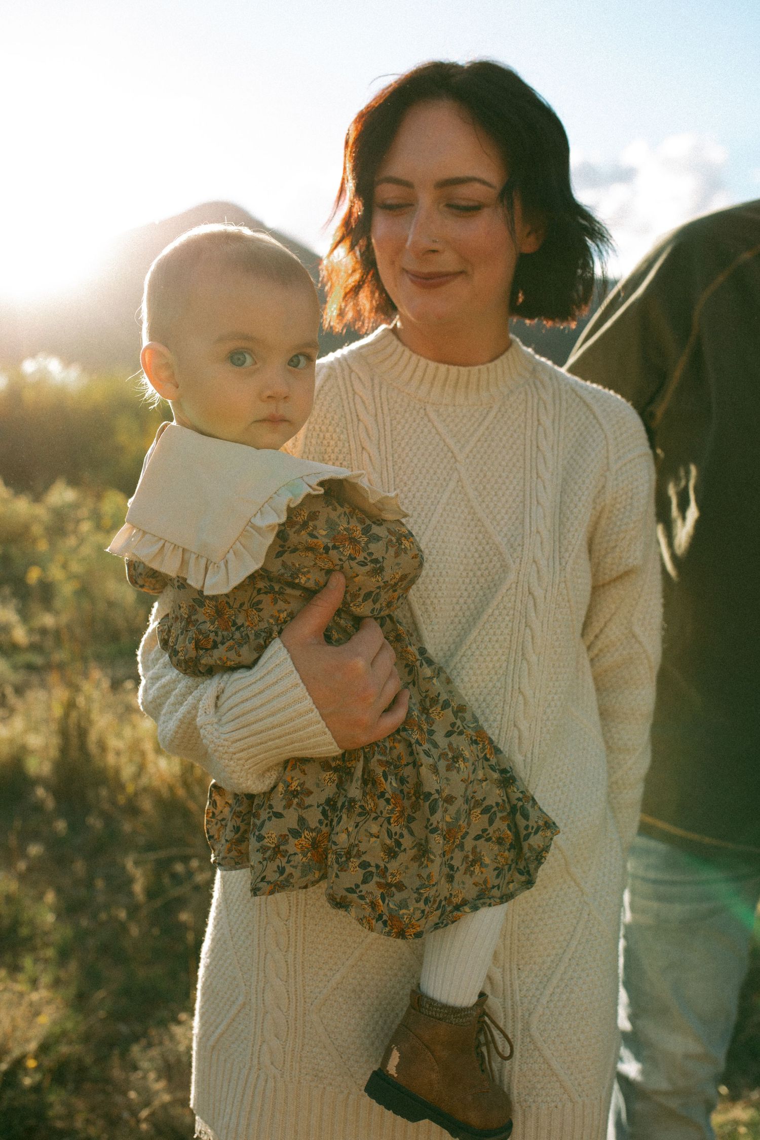 Woman in a cream cable-knit sweater dress smiles with closed eyes while holding a baby in a floral dress outdoors in golden sunlight.