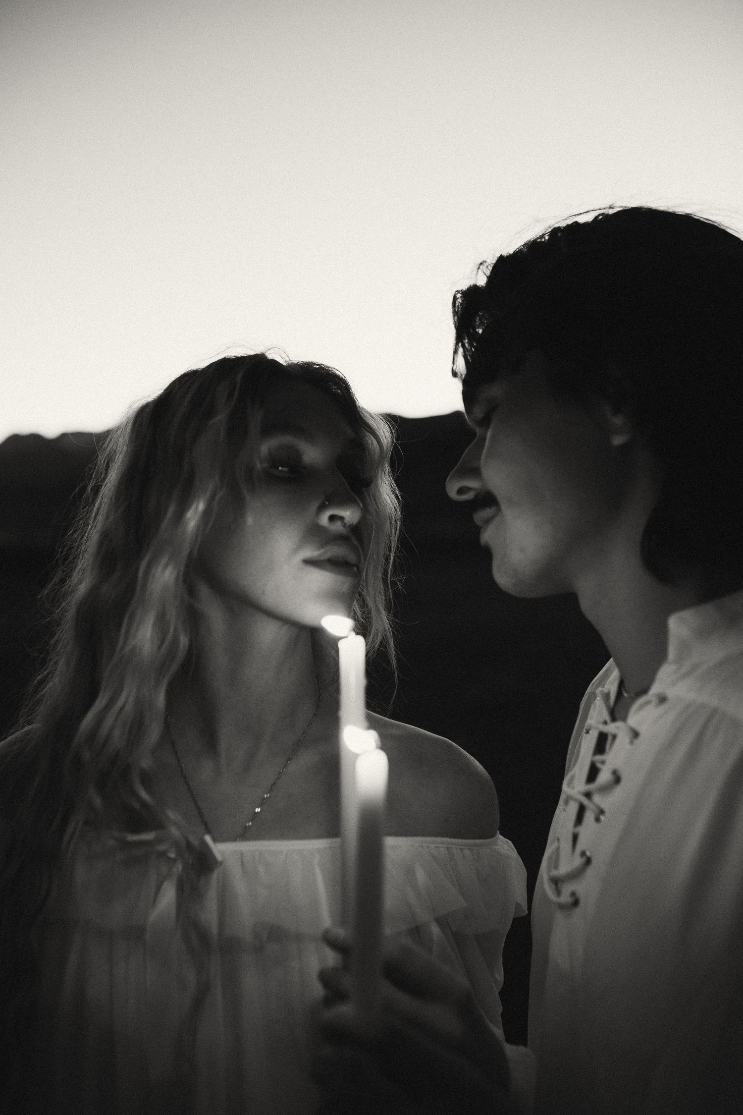 Black and white portrait of a couple sharing a quiet moment, holding lit candles close together at dusk, faces nearly touching.