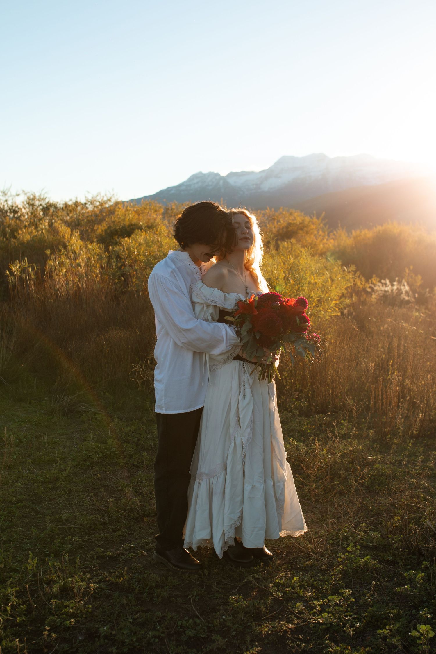 Couple embracing in a golden meadow at sunset, the bride in a white dress holding a red bouquet with snowy mountains behind them.