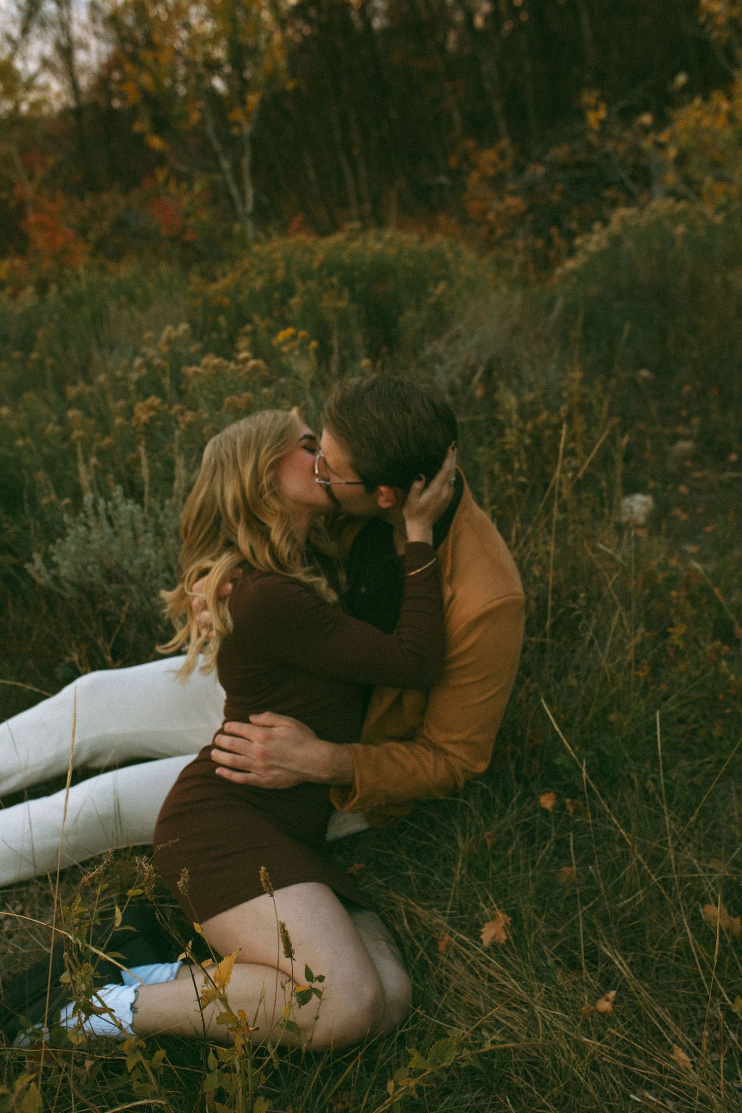 Couple kissing while sitting in a grassy autumn field, woman in brown dress and man in rust jacket, romantic engagement photo.