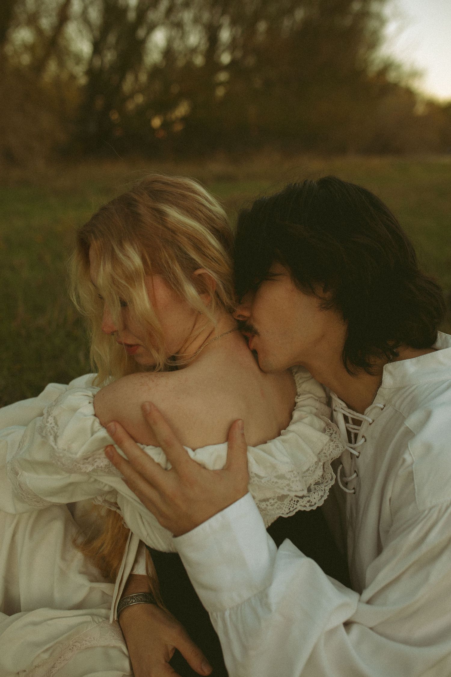 Romantic couple in white vintage-style clothing embracing in a grassy field at golden hour, his lips near her bare shoulder.
