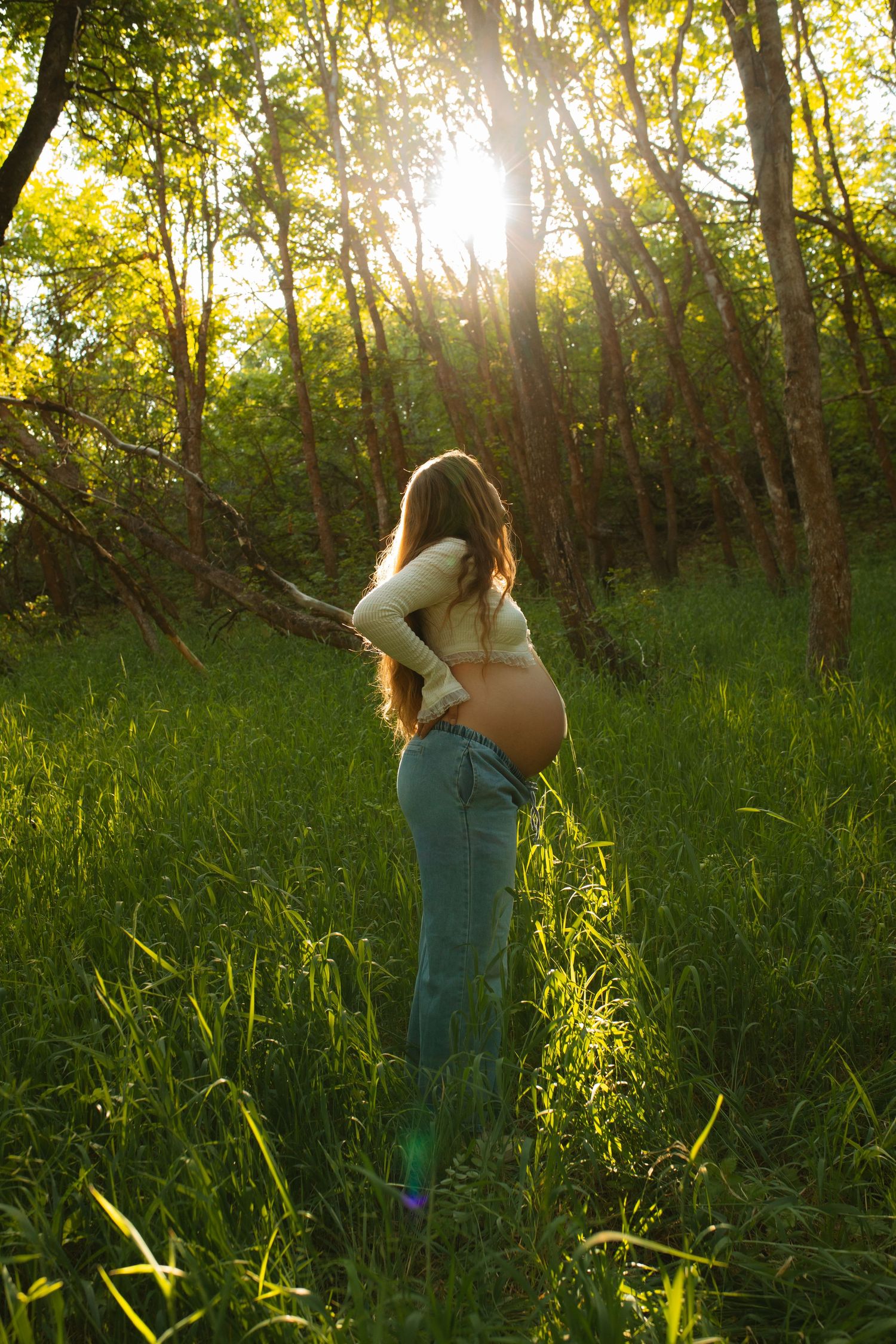 Pregnant woman with long hair stands in a sunlit green forest meadow, her bare belly glowing in golden light.
