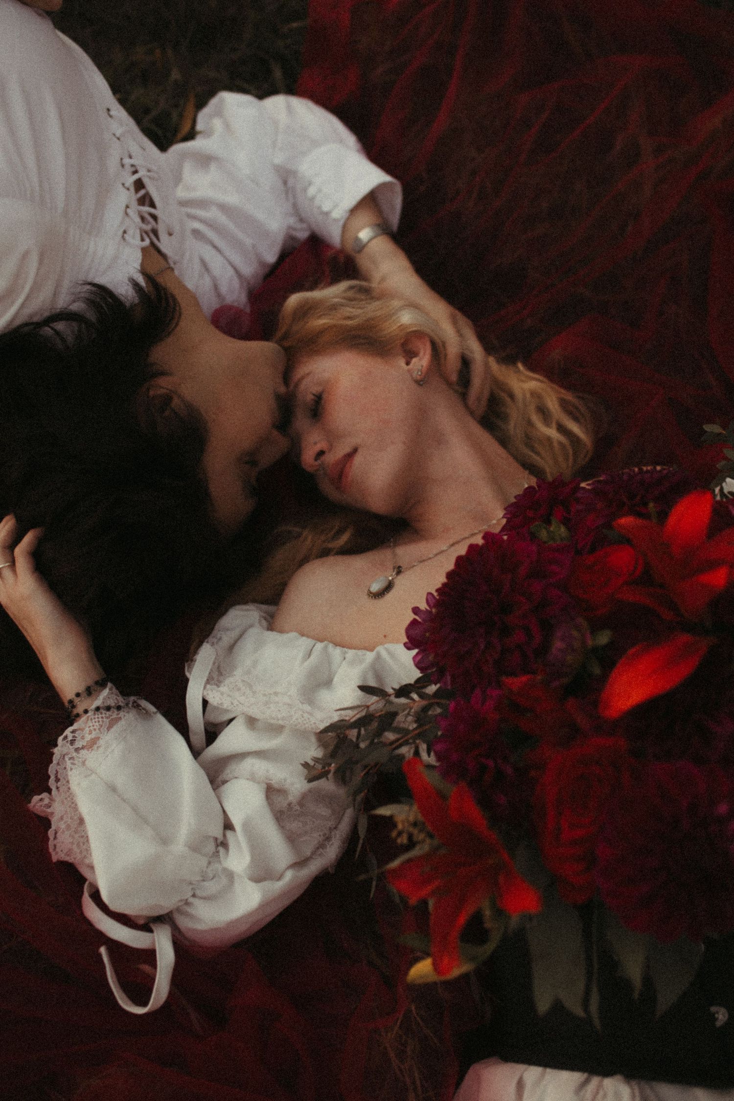 Two people in white romantic blouses lie close on a deep red velvet blanket beside a bouquet of dark red dahlias and lilies.