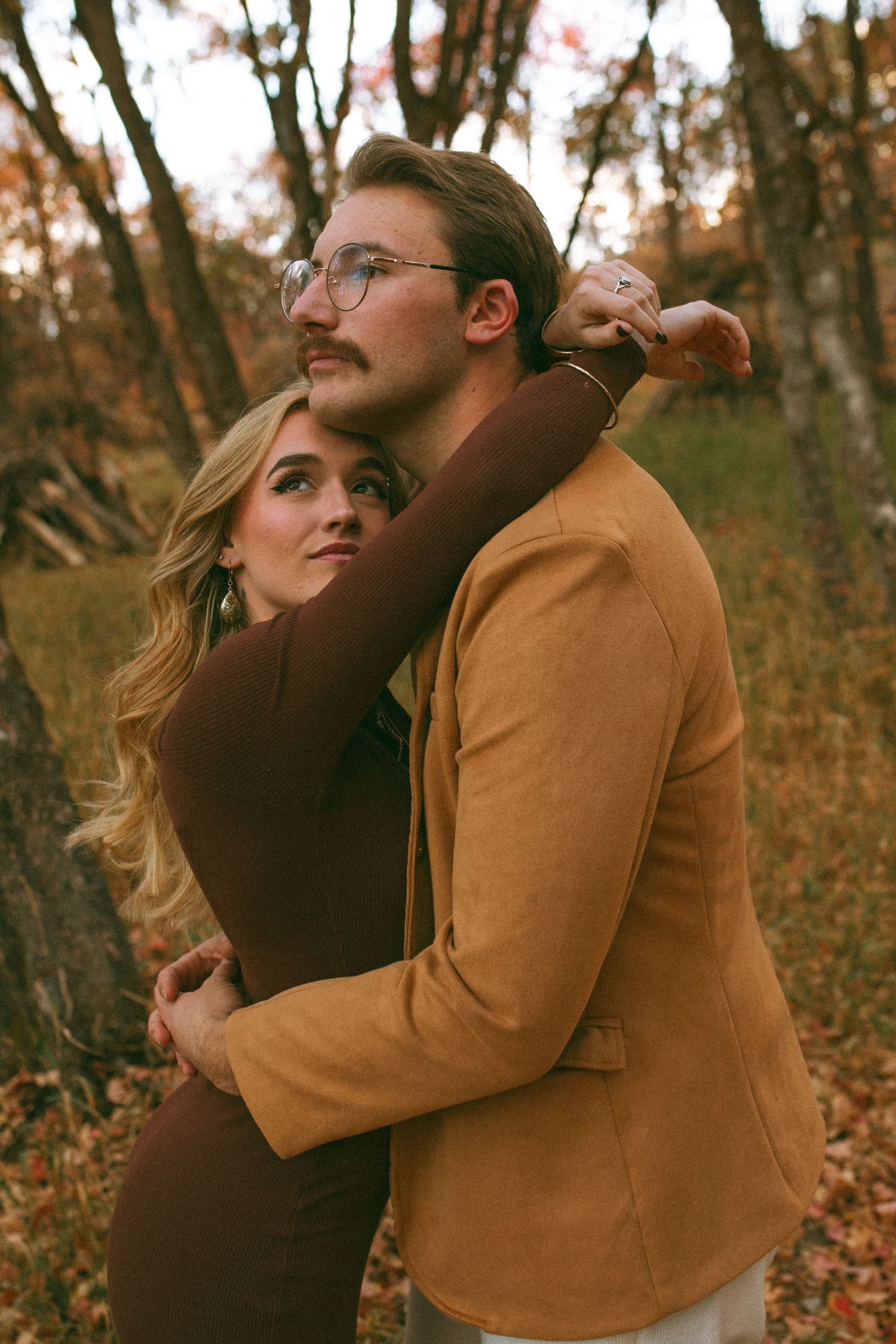 Couple embracing in an autumn forest, the woman in a brown dress hugging a bearded man in glasses and a tan suede jacket.