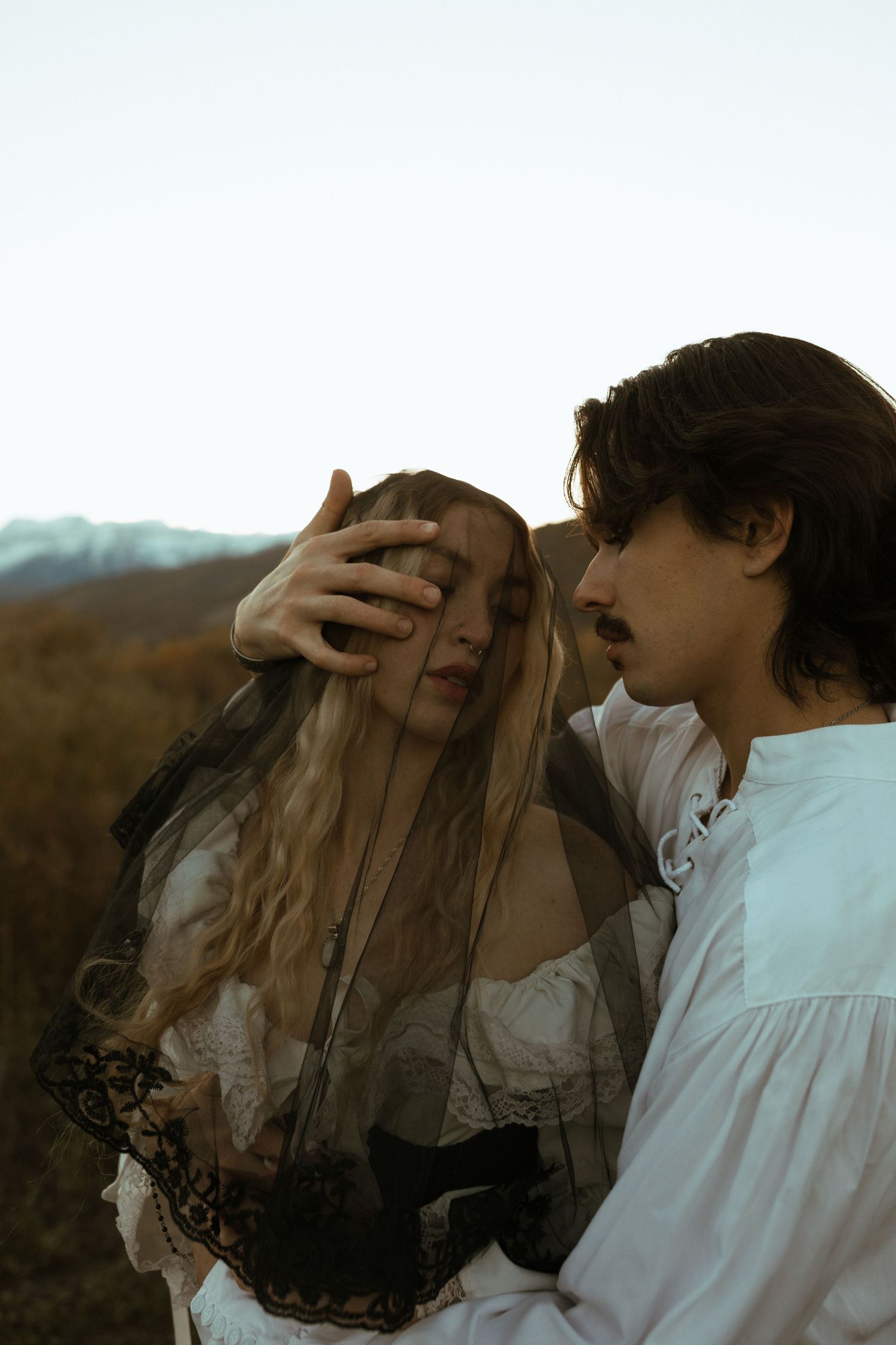 Man in a white poet shirt shields a woman's face with his hand as she wears a black lace veil, with mountains in the background.