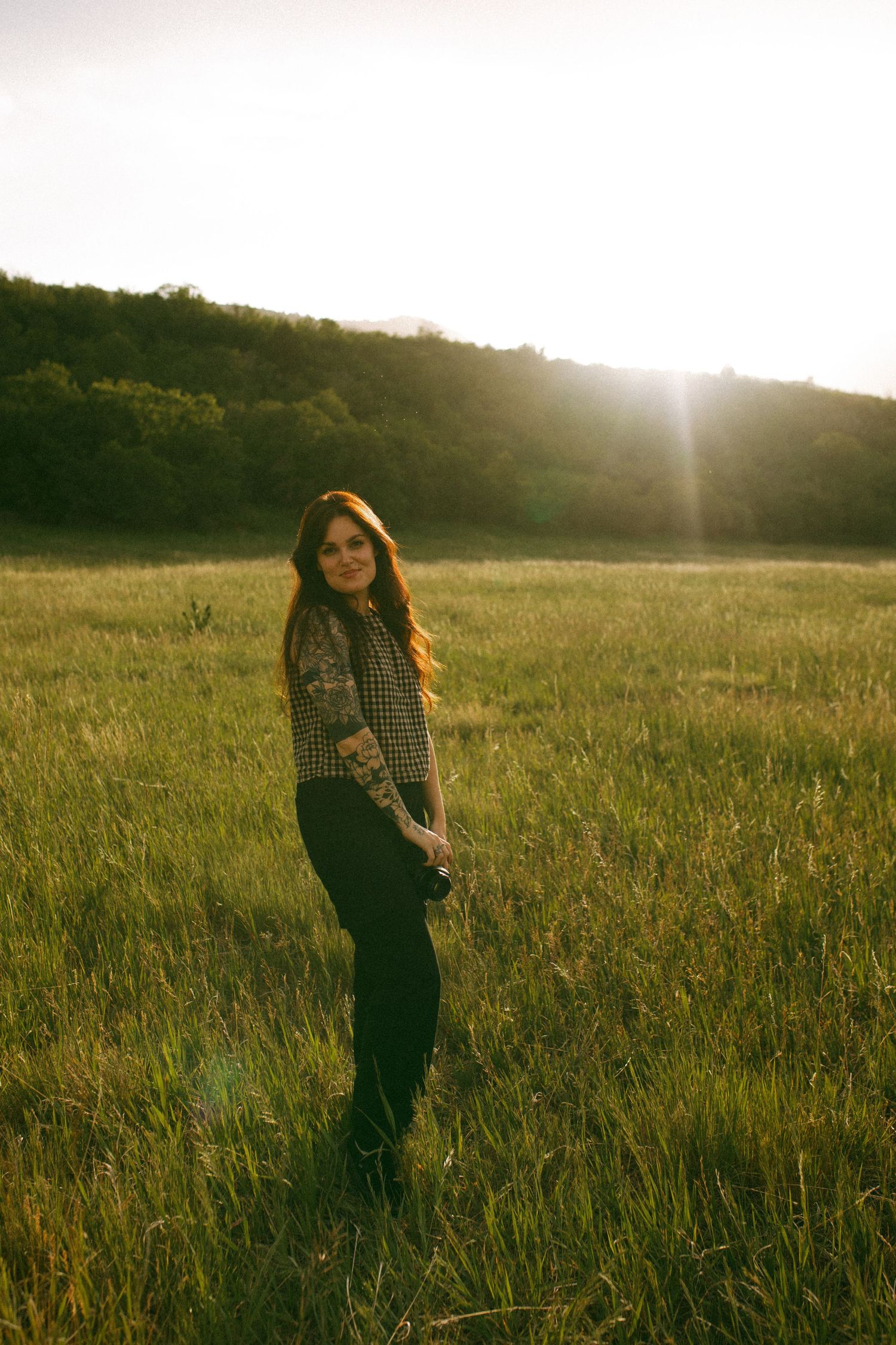 Woman with long auburn hair and tattooed arms stands in a golden grassy field at sunset, holding a camera with forested hills behind her.